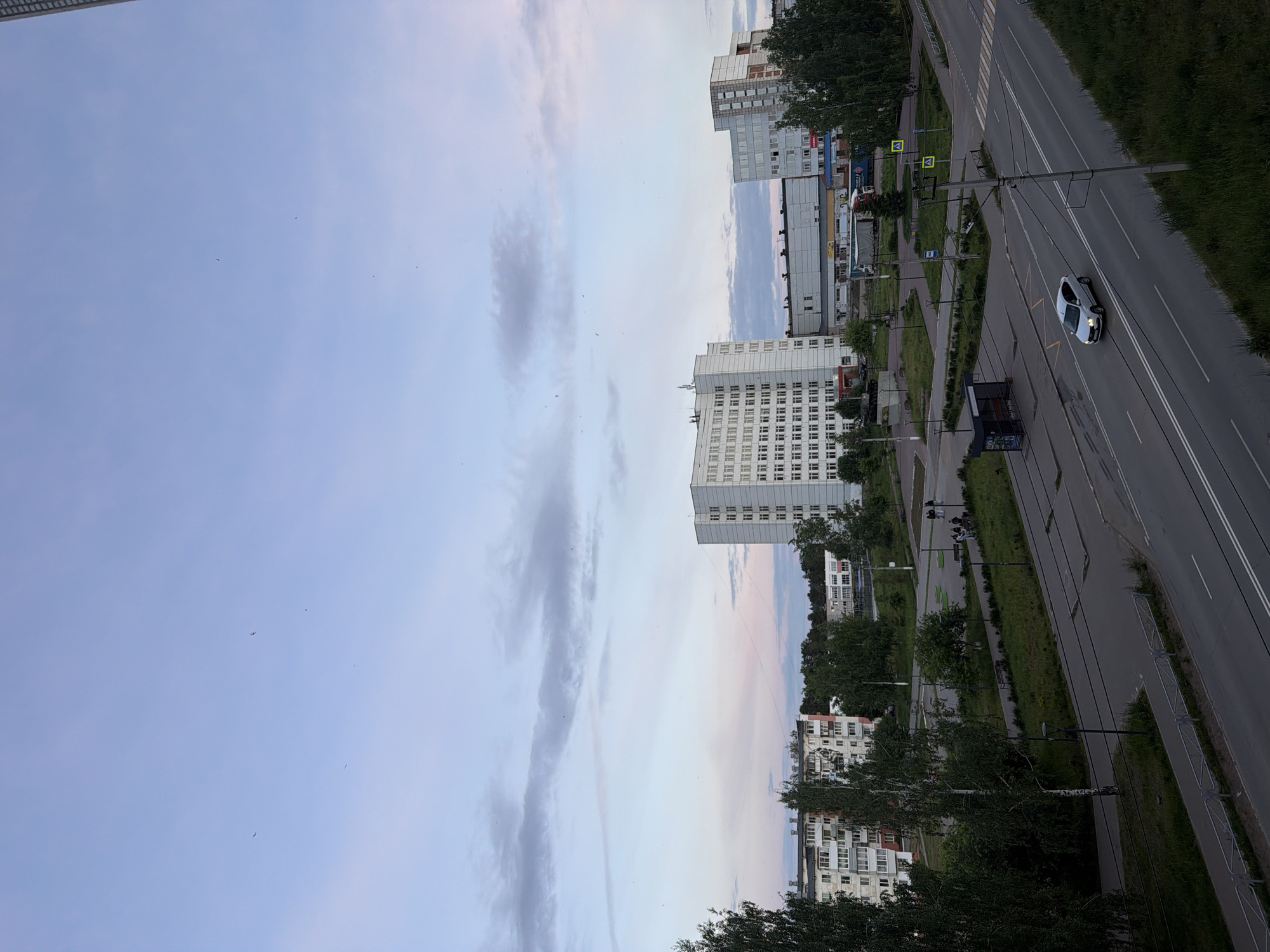 This elevated view captures an urban landscape in Chaikovsky, Russia, under a partially cloudy, light blue sky, possibly during early morning or late evening due to the soft, diffused light.
In the midground, a wide, multi-lane asphalt road extends horizontally across the frame. It features clear white lane markings and overhead power lines, likely for trolleybuses. A white sedan, with its headlights on, is driving in the rightmost lane, while a dark, boxy truck or van is visible closer to the foreground, near the roadside. Several pedestrian crossings, marked with zebra stripes on the road and yellow square signs with walking person symbols on poles, indicate pedestrian infrastructure.
To the left of the road, a series of multi-story buildings dominate the landscape. These appear to be predominantly white or light-colored residential or mixed-use structures, characterized by numerous windows and some balconies. Further in the background and to the right, more buildings of varying sizes and architectural styles can be seen.
Greenery is abundant throughout the scene, with numerous trees, including identifiable birch trees, lining the road and interspersed between the buildings. Grassy areas are also visible. Streetlights are present along the road and within the green spaces. There are no clearly visible individuals, and no legible text can be discerned on buildings, signs, or vehicles within the image. The scene conveys a sense of a quiet, functional urban area.