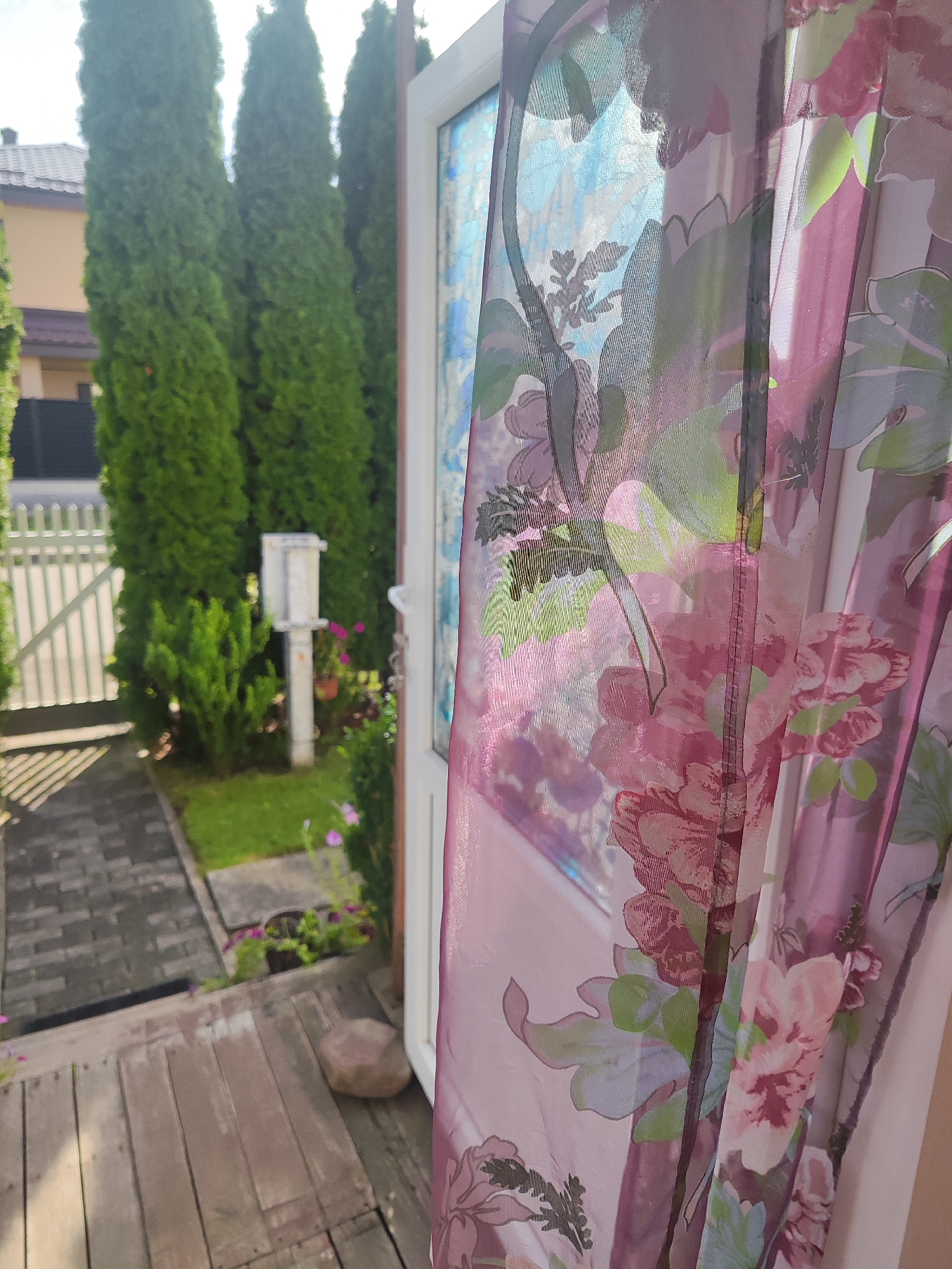 The image shows a close-up of a sheer floral curtain with pink and purple flowers, obscuring a view of a residential area. Through the translucent fabric, one can discern a doorway, tall green trees, a house with a tiled roof, a white picket fence, and a paved walkway leading to a wooden porch. The overall setting appears to be a yard or garden. The photo was taken in Gvardeysk, Russia.