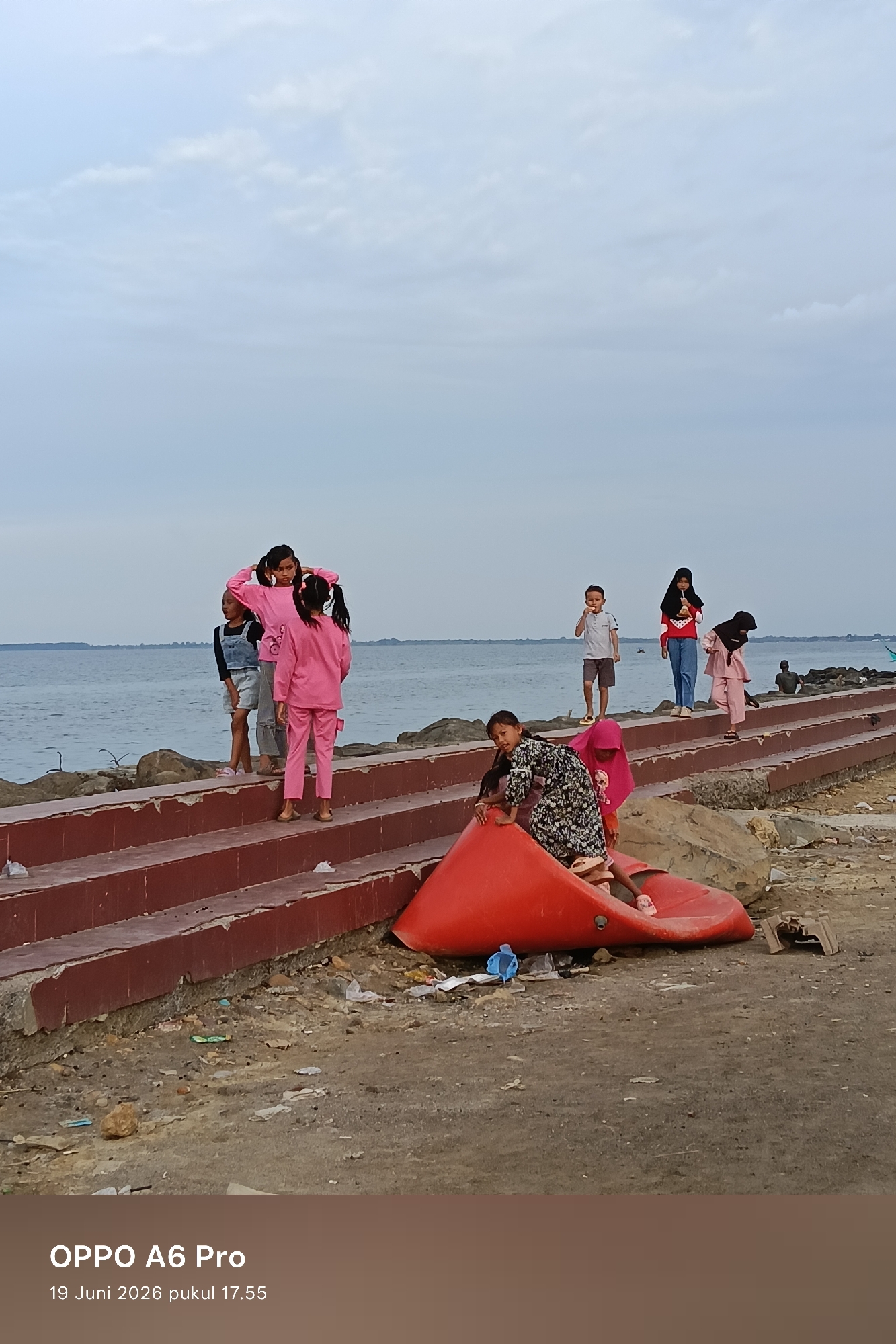 In the city of Lhokseumawe, Indonesia, on the coast, a group of children are gathered near the sea. A few are on a red concrete embankment, with a body of water to their left. The sky is overcast, and the sea appears calm. Some of the children are wearing casual clothing, while others are in traditional attire, including hijabs. One child is sitting in a large, bright red plastic structure that resembles a slide or boat. The area around the embankment is strewn with litter. In the foreground, text indicates the image was taken on "19 Juni 2026 pukul 17.55" with an "OPPO A6 Pro" phone.