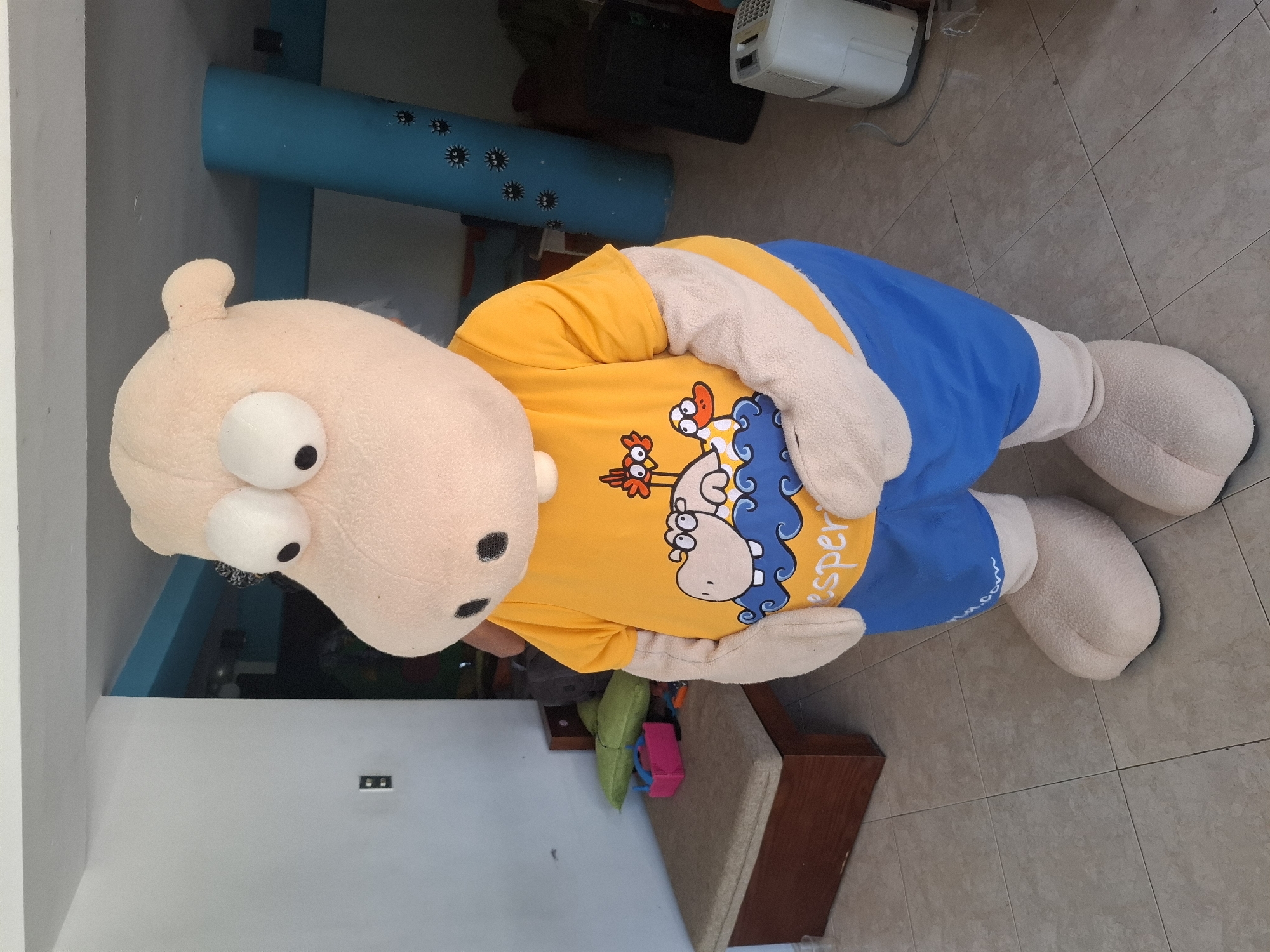 The image depicts a person in a light-colored hippopotamus mascot costume standing indoors. The mascot is wearing a bright orange t-shirt with a cartoon design featuring a hippopotamus, two ducks, and wavy blue lines. The t-shirt also has white trim. Below the t-shirt, the mascot wears blue shorts with a white trim and a white waistband. The mascot has large, bulging white eyes with black pupils, small ears, and a rounded nose. Its arms are bulky and light-colored, and its feet are also large and light-colored.
The mascot is standing on a tiled floor with a grid pattern. To the left of the mascot, there is a wooden bench with a light brown padded seat. Behind the bench, there is a light gray wall with a black electrical outlet. A bright green pillow and a pink toy are visible on the bench. In the background, a large, bright blue cylindrical object with black star-like patterns is present. Further in the background, a black electronic device and a white appliance resembling a dehumidifier are visible. The overall environment appears to be a casual indoor space, possibly a recreational area or a business. Based on the provided context, this scene is located in Playa El Agua, Venezuela.