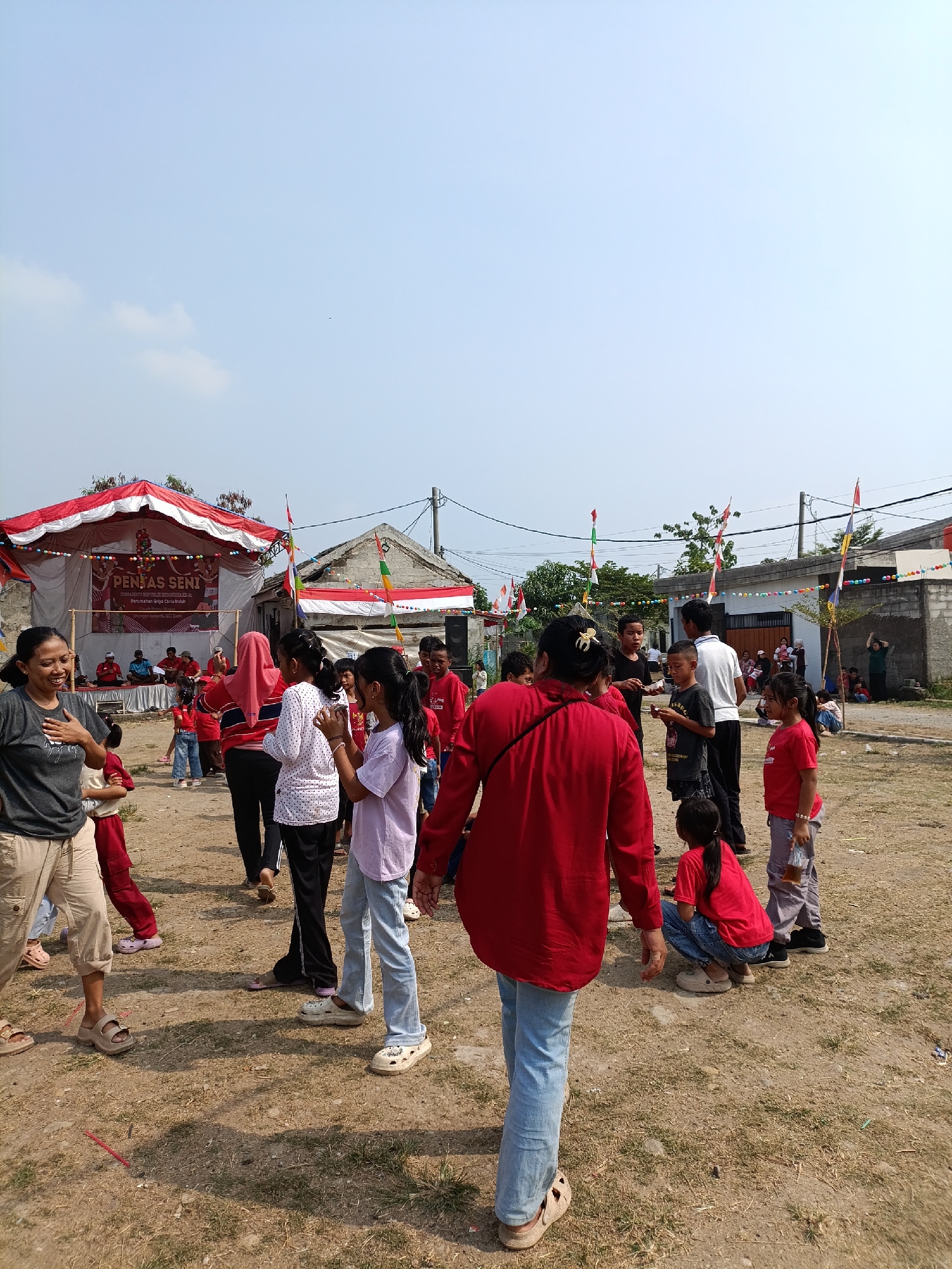 The image depicts an outdoor event in the city of Cariu, Indonesia, likely a community gathering or performance, as indicated by the stage setup in the background. A tent with red and white stripes is erected, behind which a banner displays text in Indonesian, including "PENTAS SENI" (Arts Performance) and "Perumahan Griya Ceria Indah" (Griya Ceria Indah Housing), suggesting the event is organized by or for residents of this housing complex. Several people, including adults and children, are present in the foreground and middle ground.
In the left foreground, a woman wearing a dark grey t-shirt and beige pants is smiling and has her hand on her chest, appearing to interact with someone off-camera. To her right, a child in a red outfit is partially visible. In the center, a woman in a vibrant red shirt and blue jeans is seen from behind, walking towards the right. Several children and adults are gathered around, some looking towards the stage, while others are engaged in conversations or activities.
The ground is covered in dry dirt and sparse grass, typical of an outdoor, undeveloped area. In the background, simple buildings and fences are visible, along with several Indonesian flags and colorful bunting strung across wires, indicating a festive atmosphere. The sky is clear and bright blue, suggesting it is daytime. The overall scene captures a lively community event taking place in an open, somewhat rustic setting.