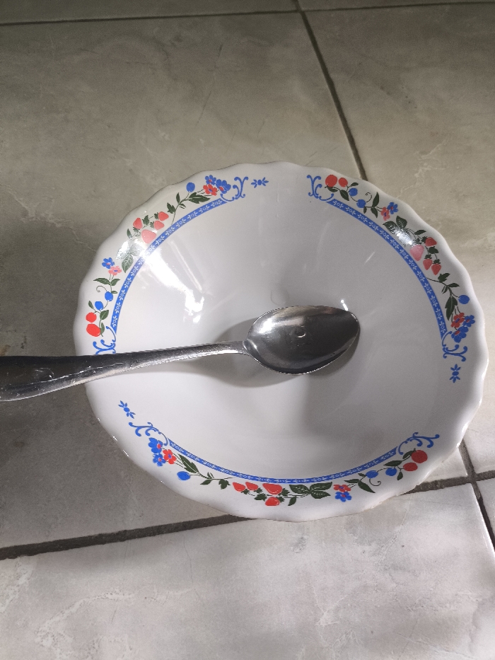 A white bowl with a scalloped edge is decorated with a blue, red, and green floral pattern. A silver spoon rests inside the bowl, casting a shadow onto the white surface. The bowl sits on a tiled surface with neutral tones. The light source is above, creating highlights and shadows on the bowl and tiles.