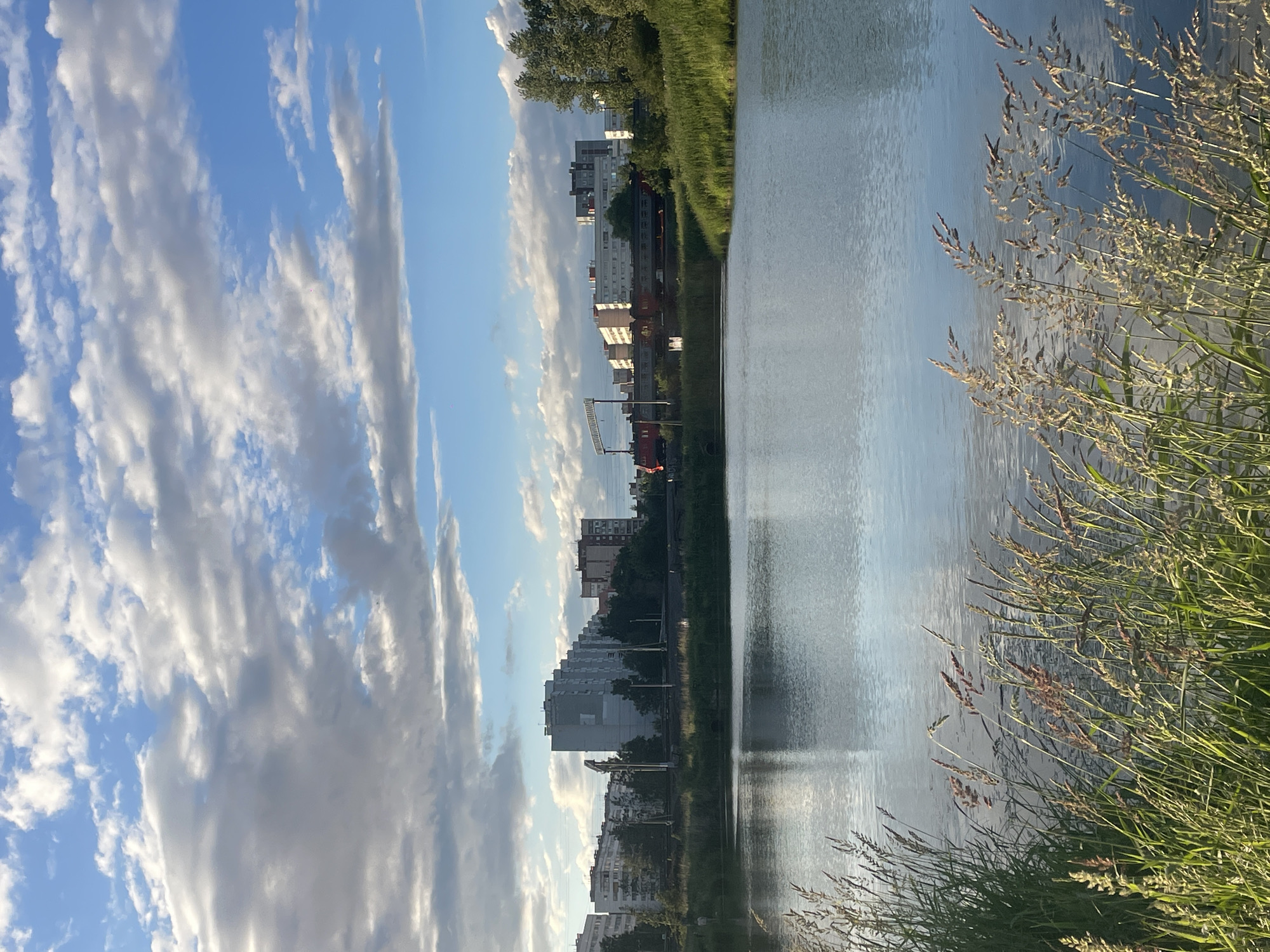 This is a vertical, eye-level shot of a body of water with an urban landscape in the background. The water is calm, reflecting the sky and clouds. In the foreground, tall grasses are visible, adding texture and depth to the scene. Across the water, a row of residential buildings is situated, with a train track and a red train in motion running in front of them. The sky is partly cloudy, with a mix of blue sky and white and gray clouds. The overall atmosphere is peaceful and serene, with a hint of urban life visible in the background.
This scene is likely located in the Nevsky district of Saint Petersburg, Russia, possibly near a park or a suburban area with a body of water and residential buildings. The presence of a train track and buildings suggests an urban environment, while the water and greenery indicate a natural element within the city.
