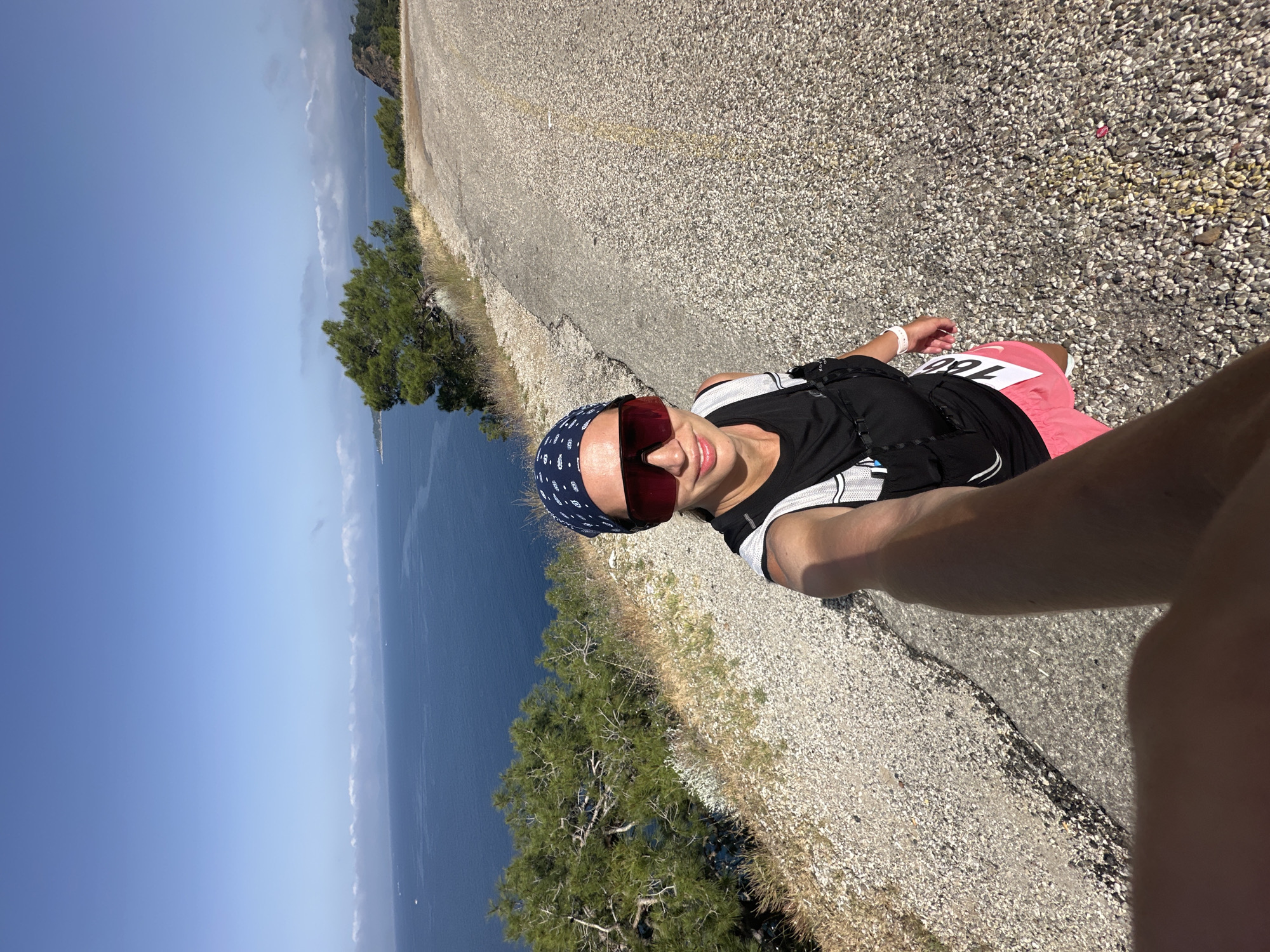 A medium shot shows a woman in athletic attire taking a selfie on a coastal road in Fethiye, Turkey. She wears a dark top with white stripes, red shorts, and a blue patterned bandana. She has sunglasses on and is holding a selfie stick. A race number, "160," is pinned to her shorts. The asphalt road is on the right side of the frame, leading toward the foreground. A steep drop to the left features a blue sea and sky. Sparse vegetation clings to the cliffside between the road and the sea.