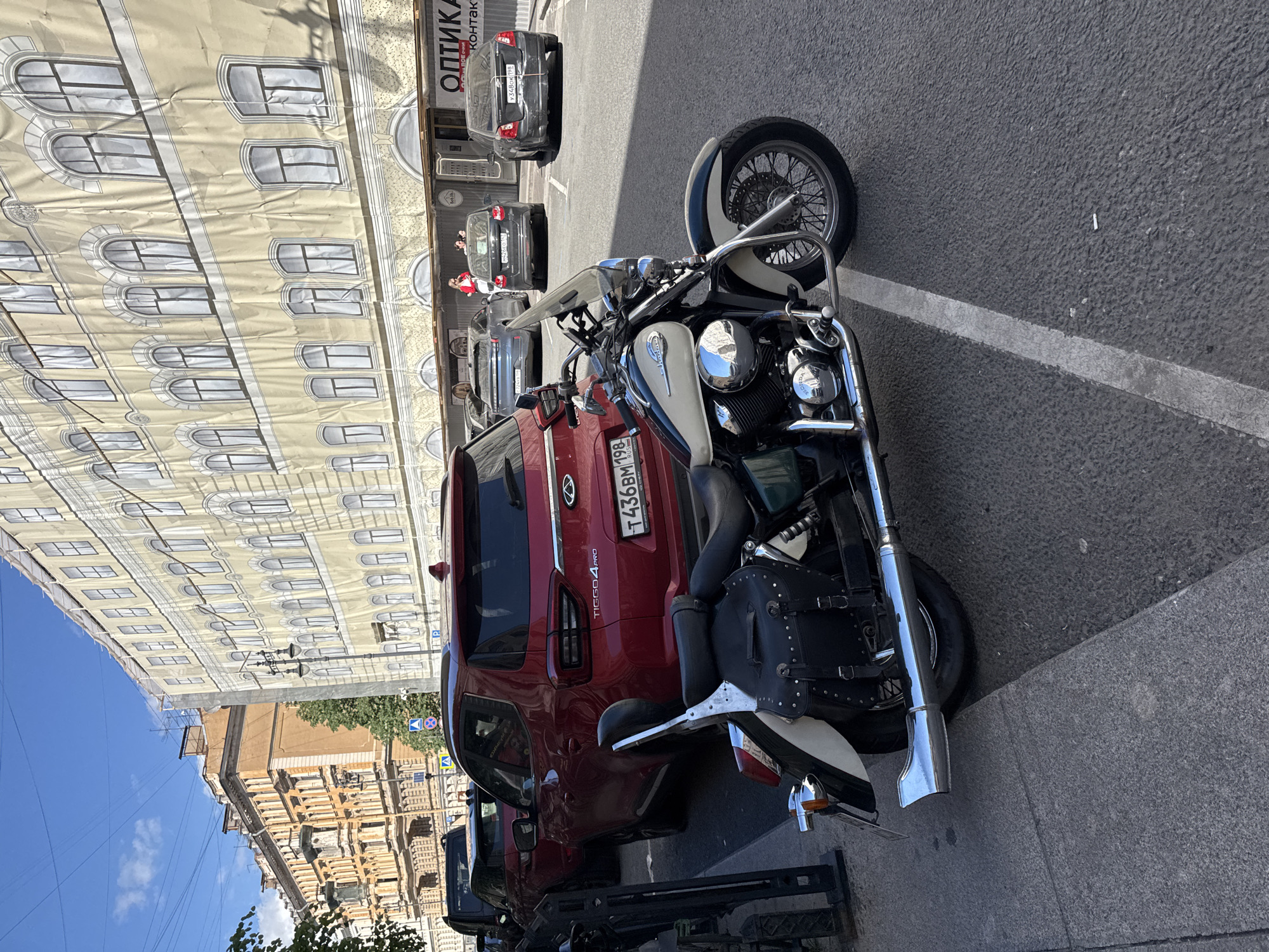 In the city of Saint Petersburg, Russia, on a sunny day, a motorcycle is parked on a paved street in front of a building with scaffolding covering its facade. The motorcycle, a cruiser style with a windshield and saddlebags, is the foreground subject. Behind it, a red SUV is parked, its license plate visible. Several other cars are parked further down the street, and pedestrians are visible near the building's entrance. The street appears to be in an urban area, with architectural elements of old buildings visible on the left. A discarded cigarette butt is on the asphalt near the motorcycle's front wheel.