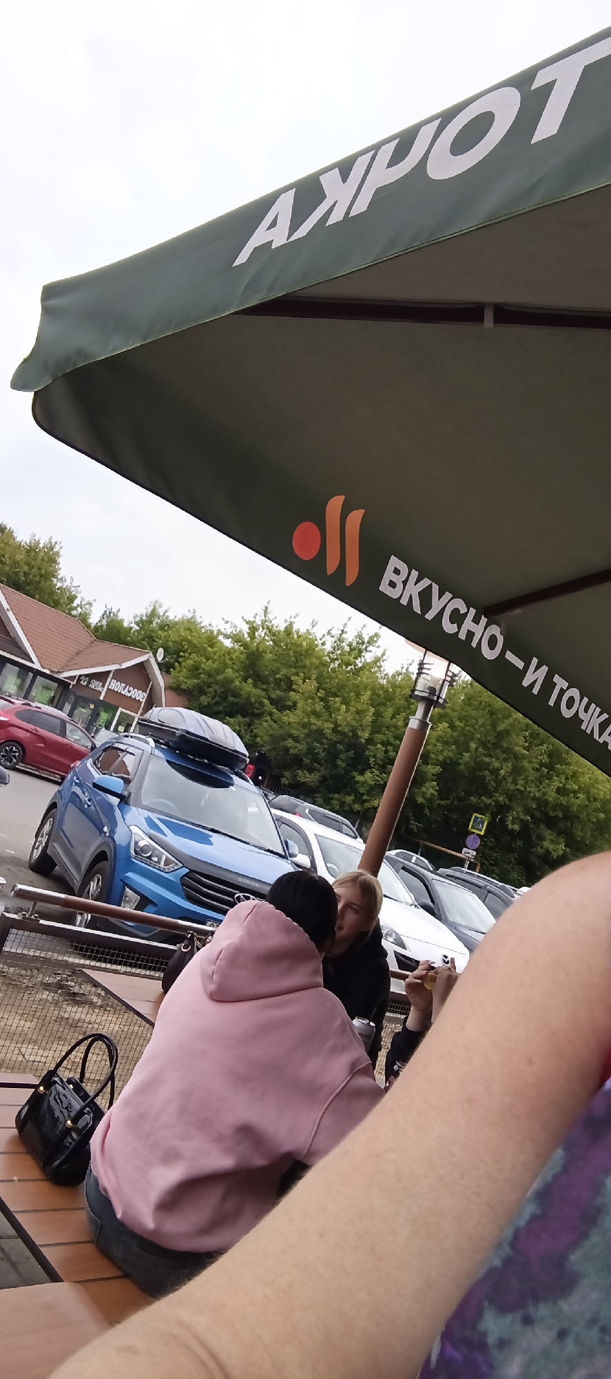 This is an outdoor, eye-level, full shot taken on an overcast day in Golitsyno, Russia. The scene is set at a cafe with an umbrella providing shade. In the foreground, a person wearing a pink hoodie sits with their back to the camera, facing a table. To their right, another person, a blonde woman, is also seated at the table. She appears to be engaged in conversation, looking towards the person in the pink hoodie, and is holding a small object, possibly a fork with food. A black patent leather handbag is placed on the table next to the person in the pink hoodie.
In the midground, a blue Hyundai Creta SUV is parked, equipped with a roof cargo box. Other cars are visible in the parking lot behind it, including a white vehicle and a dark sedan. The cafe building has a distinctive brown tiled roof and a sign with Cyrillic text that appears to read "ВОЛКАРД" in reverse.
The umbrella overhead features white text that reads "АХРОТ" and "ВКУСНО - И ТОЧКА". The "ВКУСНО - И ТОЧКА" portion is accompanied by an orange logo with two abstract shapes. The presence of the cafe, cars, and people suggests a casual dining or resting experience. The overcast sky indicates it is likely daytime, but not sunny.