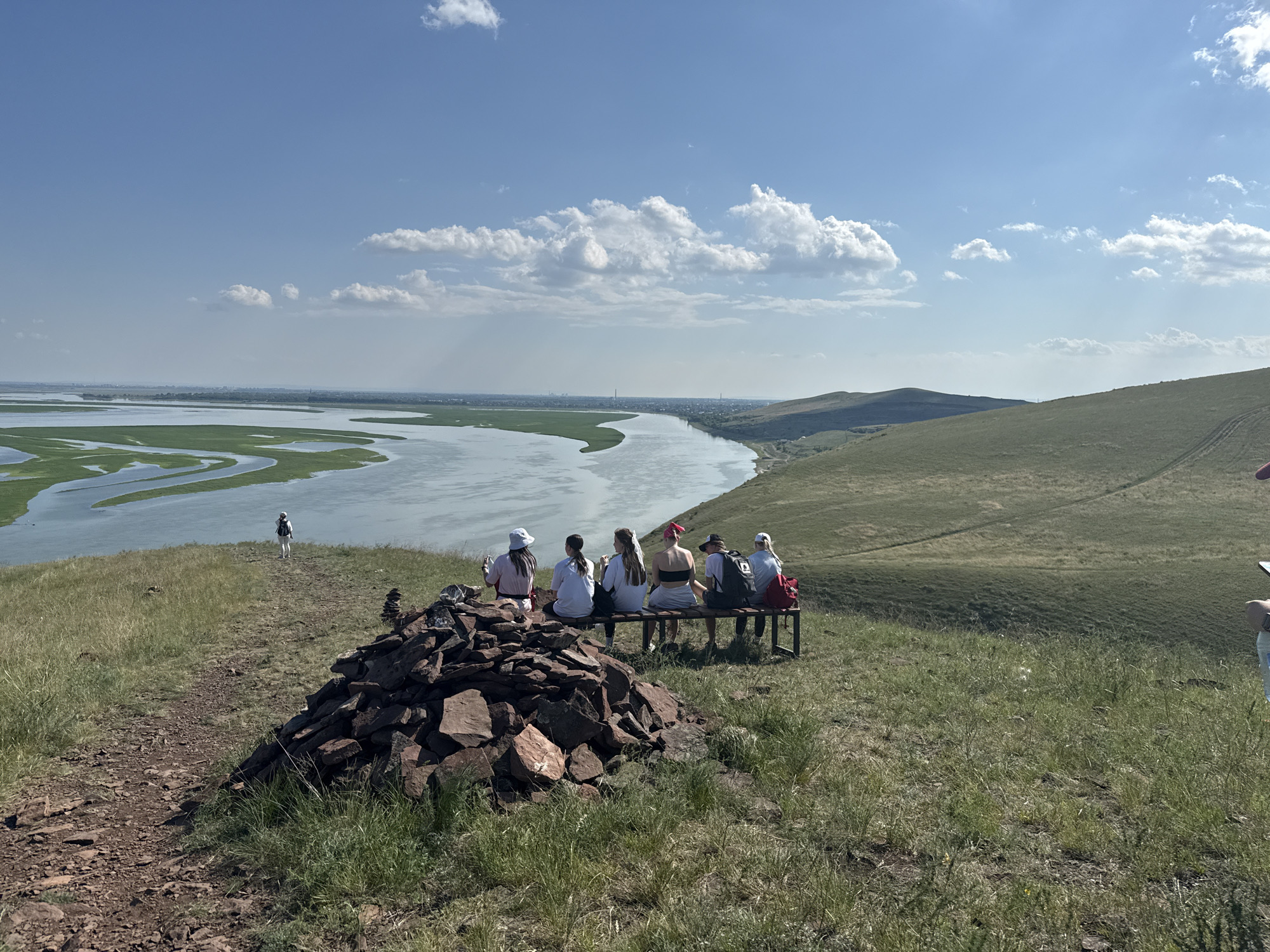 A group of six people are seated on a metal bench on a grassy hill overlooking a wide river with green islands. A pile of red rocks is in the foreground. In the distance, a lone person walks on a path. The sky is blue with scattered clouds. The location is on a hill near the city of Ust'-Abakan, Russia.