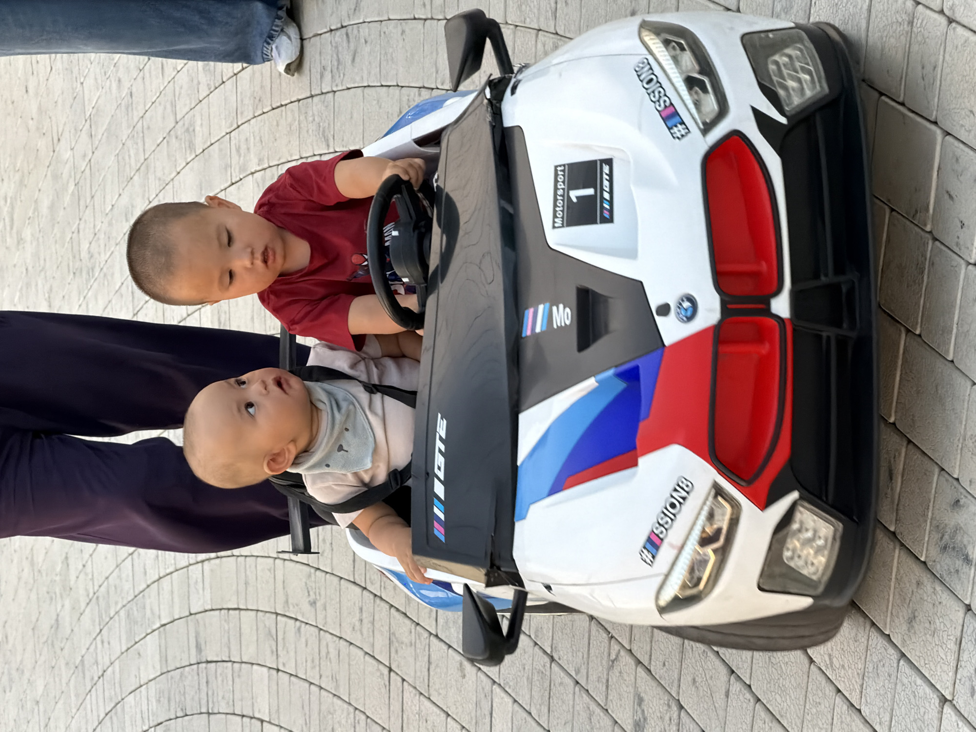 Two young children, likely toddlers, are seated in a white electric toy car designed to resemble a BMW M-series vehicle. The car is white with blue, red, and black racing stripes. The child on the left wears a light pink shirt and a light blue bandana with a cartoon animal face. The child on the right wears a red t-shirt and is positioned behind the steering wheel. The toy car has visible headlights, a grille, and side mirrors. The children are outdoors, with a paved walkway made of light-colored interlocking bricks visible in the background. The lower halves of two adults' legs are visible, one on the left wearing dark purple pants and the other on the right wearing blue jeans. The scene appears to be a public outdoor space in a city.
