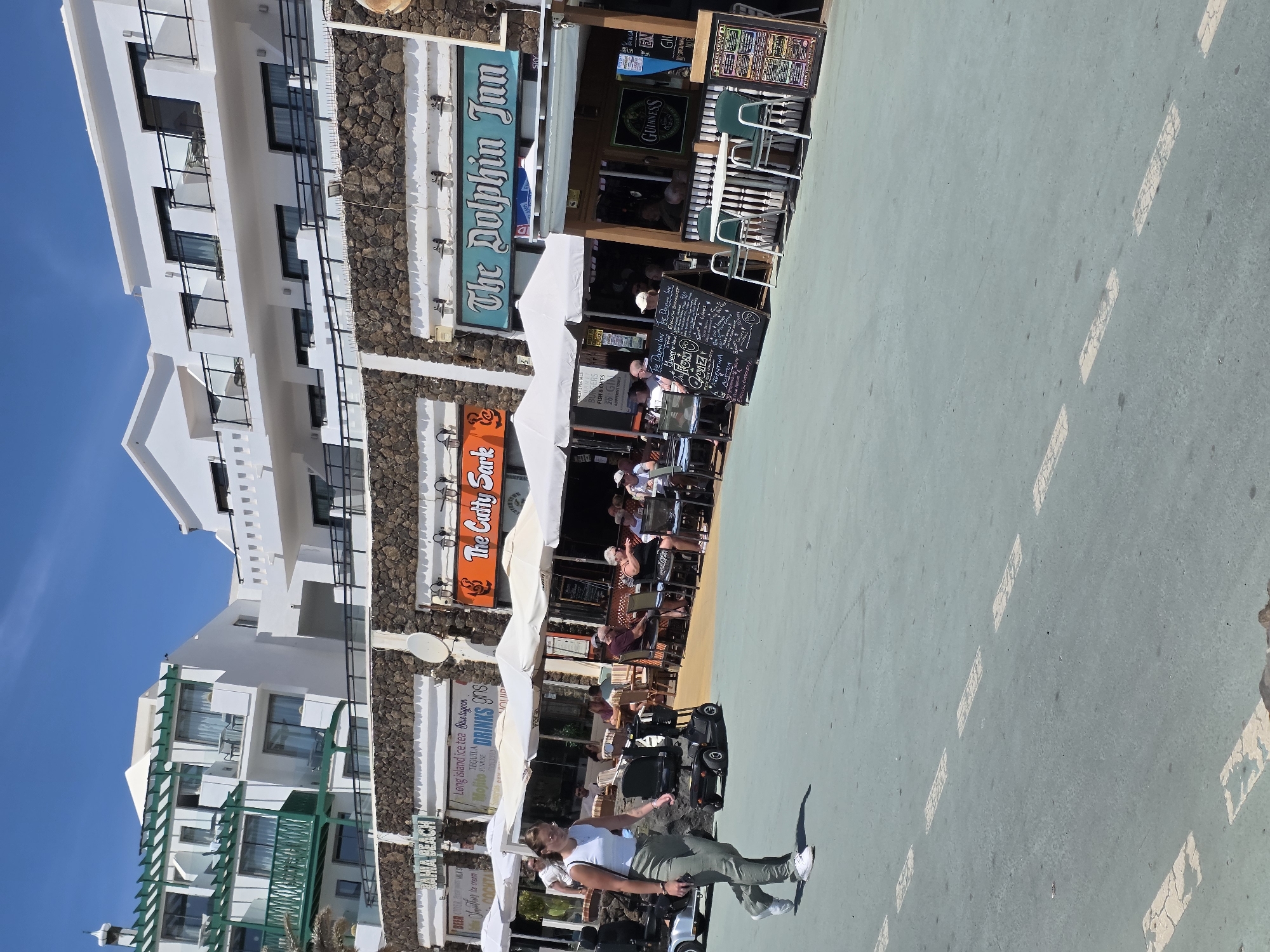 A woman in a white tank top and green pants walks left to right in front of several outdoor cafes. She is wearing sunglasses and white sneakers. In the background, there are white buildings with balconies and a stone facade. Two mobility scooters are parked on the left side of the image. The cafes have white umbrellas and tables with people seated. Signs are visible above the cafes, including "BAHIA BEACH", "The Cutty Sark", and "The Dolphin Inn". The ground is a light blue-green color with white dashed lines. The sky is clear and blue. This scene is likely in Teguise, Spain, given the architecture and ambiance common to tourist areas in the Canary Islands.
