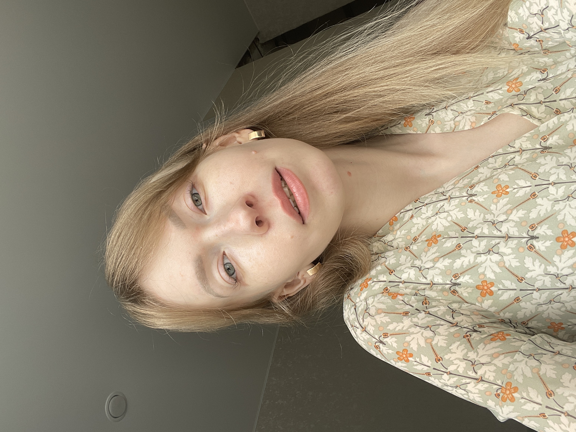 A medium shot depicts a fair-skinned individual with long, blonde hair, wearing gold, rectangular earrings and a light green patterned top. The person is positioned at an angle, with their face closer to the camera. The background consists of a smooth, gray surface, possibly a wall. The light is soft and diffused. The photograph appears to be taken indoors. The information about the location in the city of Perm, Russia cannot be confirmed with the visual data.