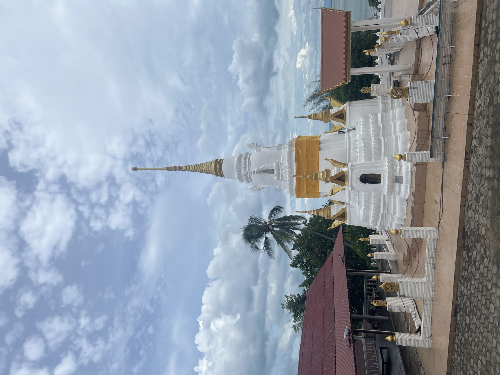 The image depicts a white stupa, a dome-shaped Buddhist monument, with gold accents and a yellow sash wrapped around its base. The stupa is tiered and has a pointed spire. It is situated within a temple complex, evidenced by the surrounding structures with red roofs and ornate golden decorations. In the background, lush green trees and a palm tree are visible, suggesting a tropical location. The sky is a mix of blue and white clouds, indicating it could be daytime with some cloud cover. The overall scene appears to be serene and spiritual. The image is oriented vertically.