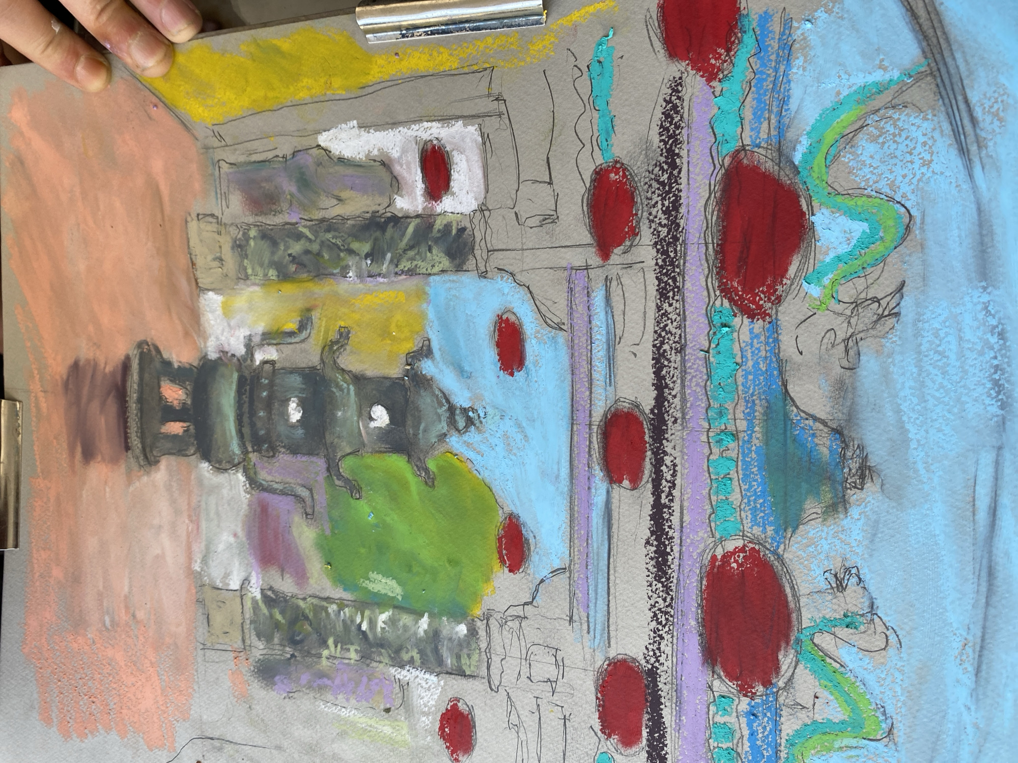 The image is a pastel drawing on gray paper, depicting an abstract interpretation of a structure, possibly a pagoda or a tiered monument. The central element is a dark, multi-layered structure with a lantern-like top. It is surrounded by vertical, column-like shapes in various shades of gray, green, and purple. Blocks of color are applied around these forms, including a large patch of bright green, light blue, and a band of pink and orange at the top. Scattered red oval shapes are present in the foreground and midground. The drawing style is loose and expressive, with visible sketch lines and smudges of pastel. The background is the gray paper, with some yellow applied to the right side. The drawing is held by a metal clip at the top and a hand is visible on the upper right edge. The setting is not identifiable as Budva, Montenegro, or any specific city.