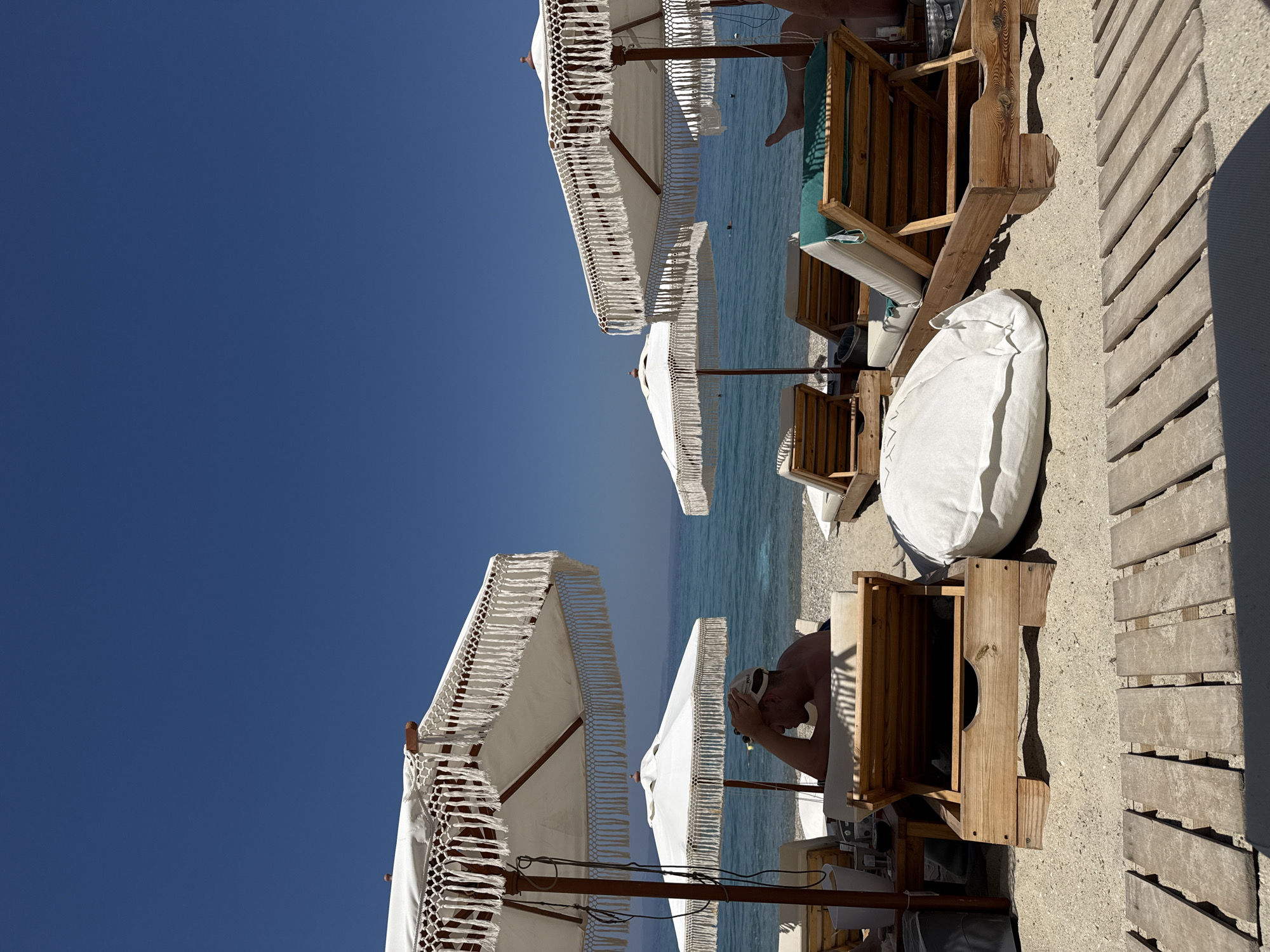The image depicts a beach scene with a clear blue sky and calm blue water. Several white beach umbrellas with decorative fringe are set up on the sand. In the foreground, a wooden lounge chair with a green cushion is visible, along with a white beanbag. A person wearing a white cap is lying on a lounge chair in the background, with their hands clasped behind their head. The lighting suggests it is daytime, likely late morning or early afternoon, with the sun casting shadows on the sand and wooden decking. The setting appears to be a beach resort or a private beach area in Pallini Municipal Unit, Greece, given the context. The overall mood seems relaxed and tranquil.