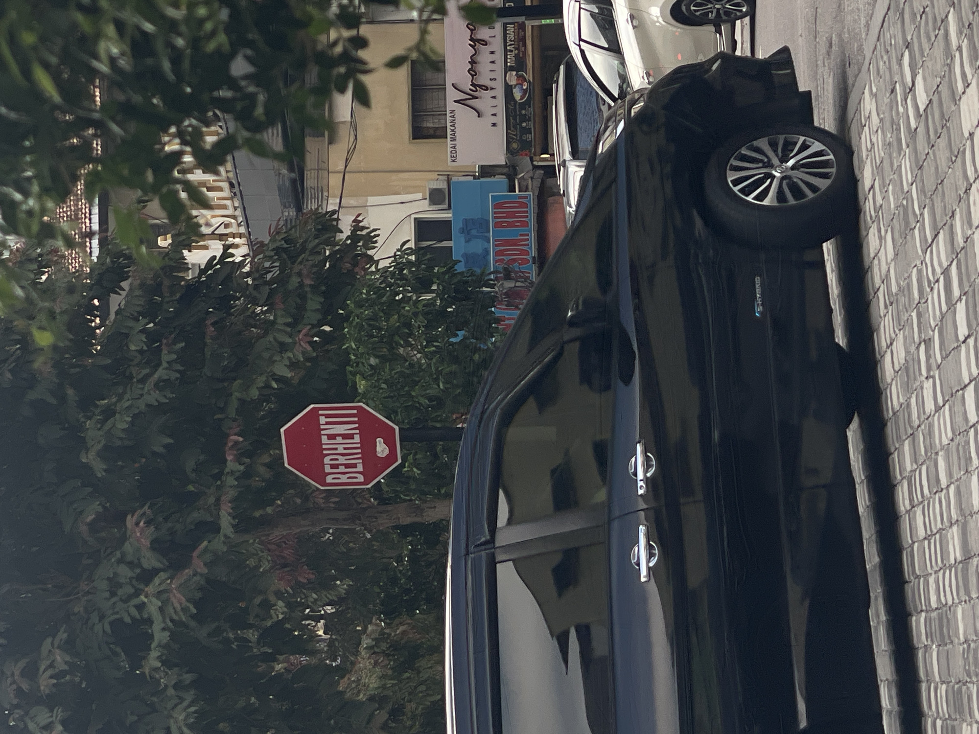 The image depicts a street scene in Kuala Lumpur, Malaysia, likely in an urban commercial area. In the foreground, the side of a black car, possibly a minivan, is prominently visible. The car has chrome door handles and a visible "e-HYBRID" badge on its lower body. It is parked on a patterned cobblestone road.
Behind the car, a red octagonal stop sign with the word "BERHENTI" (which means "STOP" in Malay) is mounted on a pole. Further in the background, several buildings are visible, some with signage. One building has a sign that reads "NYONYA MALAYSIAN CUISINE". Another sign in blue reads "SDN. BHD.", a common abbreviation for Sendirian Berhad, a type of private limited company in Malaysia. Trees with dense foliage partially obscure the buildings. The overall impression is a busy street with traffic and commercial establishments.