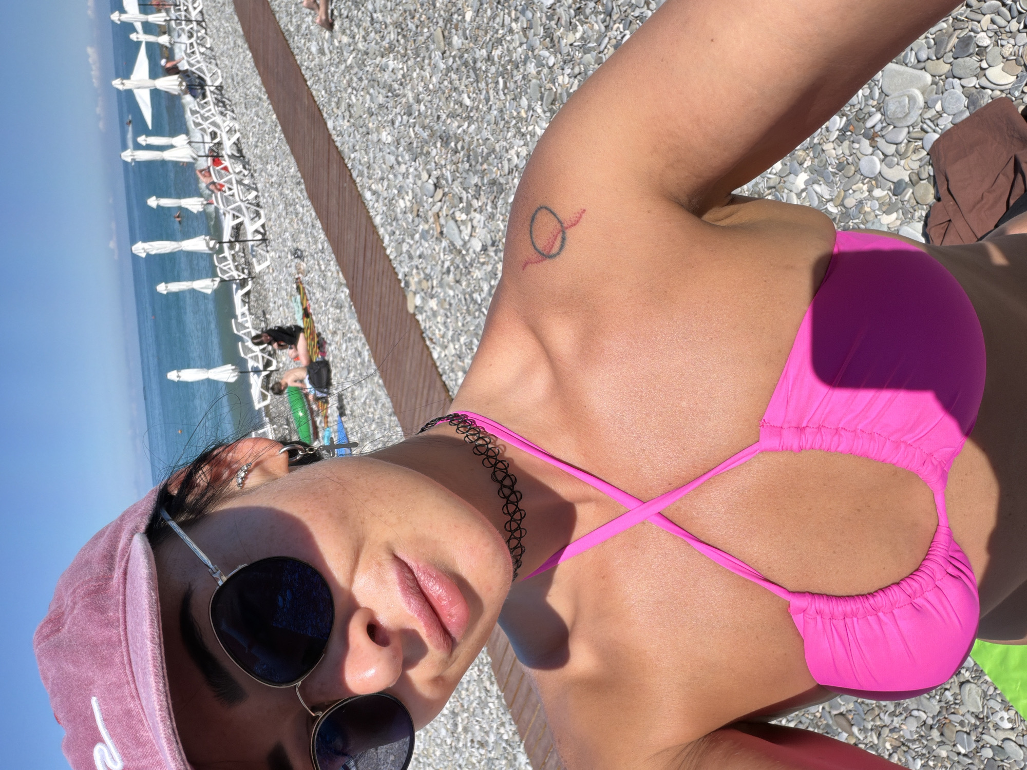 A woman in a pink bikini and a pink baseball cap is taking a selfie on a pebble beach. The ocean and a clear blue sky are visible in the background, along with rows of white beach chairs and umbrellas. She is wearing sunglasses and a black choker necklace. There is a small tattoo of a circle with a red line on her shoulder.