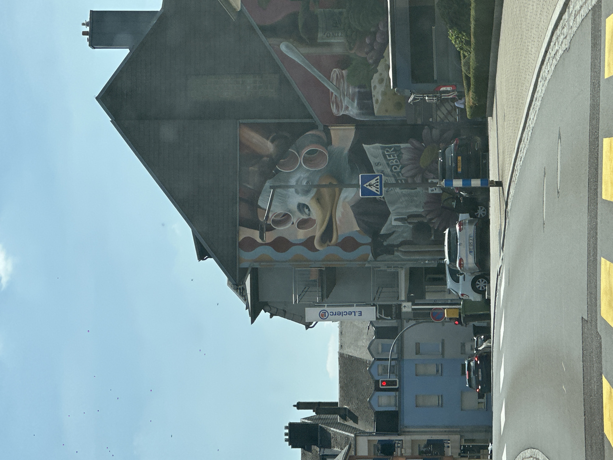 The image depicts a street scene in Käerjeng, Luxembourg, on a sunny day. A large, colorful mural featuring a cartoonish bird-like character with multiple pairs of glasses and a jar of jam is painted on the side of a building. In front of the mural, a blue and white striped pole stands next to a pedestrian crossing sign. Several cars are visible, including a white SUV and a black SUV parked on the street. Adjacent to the mural building is a blue and gray residential building with a red traffic light hanging above the street. A white sign with "E. Leclerc" and a red circular logo is attached to the side of one of the buildings. The street in the foreground has yellow and white crosswalk markings. In the sky, there are several small, dark specks, possibly birds or balloons.