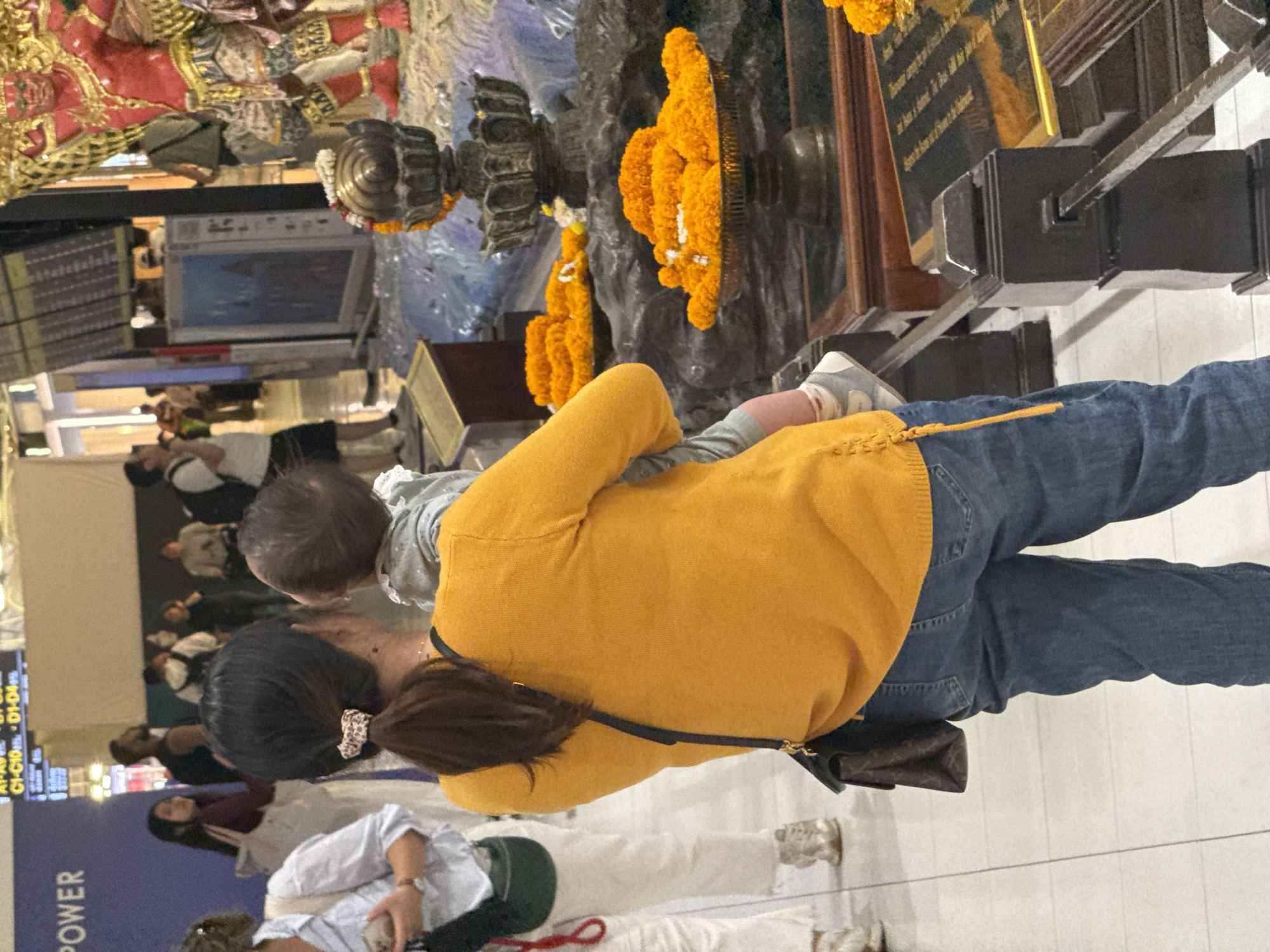 The image depicts a scene with several people in what appears to be an indoor public space, possibly an airport or a shopping mall, given the presence of a large departure board displaying text. The primary focus is on a woman wearing a bright yellow sweater and blue jeans, holding a baby. The baby, dressed in a grey outfit, is facing away from the camera. The woman has her hair tied back with a patterned scrunchie and is wearing a small, dark bag slung across her body.
In the background, other individuals are visible, some standing and others seated. There are also decorative elements, including large orange floral arrangements, possibly marigolds, which are characteristic of offerings in some Southeast Asian cultures. Behind these arrangements, a dark, sculpted object is visible, along with what looks like a sign or plaque with text.
The lighting suggests it is daytime, and the indoor setting implies a controlled environment, so weather is not a factor. The overall mood appears neutral, with no strong indicators of specific emotions. The woman's posture and the way she is holding the baby suggest a nurturing and attentive presence.
Visible text includes "POWER" on a blue surface in the lower left corner, and some unreadable text on the departure board and the sign in the background. The location context provided places the scene in Nong Prue Subdistrict, Thailand, which aligns with the cultural elements observed.