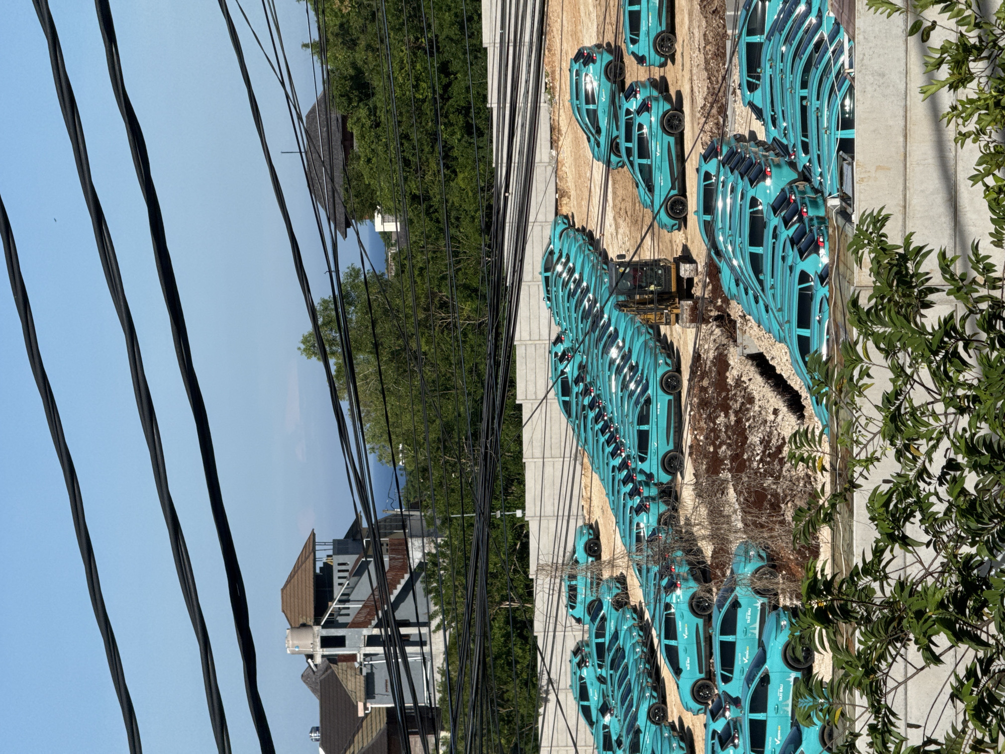 The image shows a parking lot filled with many aqua-colored cars, likely taxis, lined up in rows in Kuta Selatan, Indonesia. The cars are parked along a light-colored concrete wall. Between the rows of cars, there is brown dirt and gravel. An earth-moving machine is visible within one of the rows of cars. In the background, there is a view of buildings and trees. Power lines stretch across the foreground, obscuring some of the view. Some foliage is visible in the lower right corner. The sky is clear and blue.