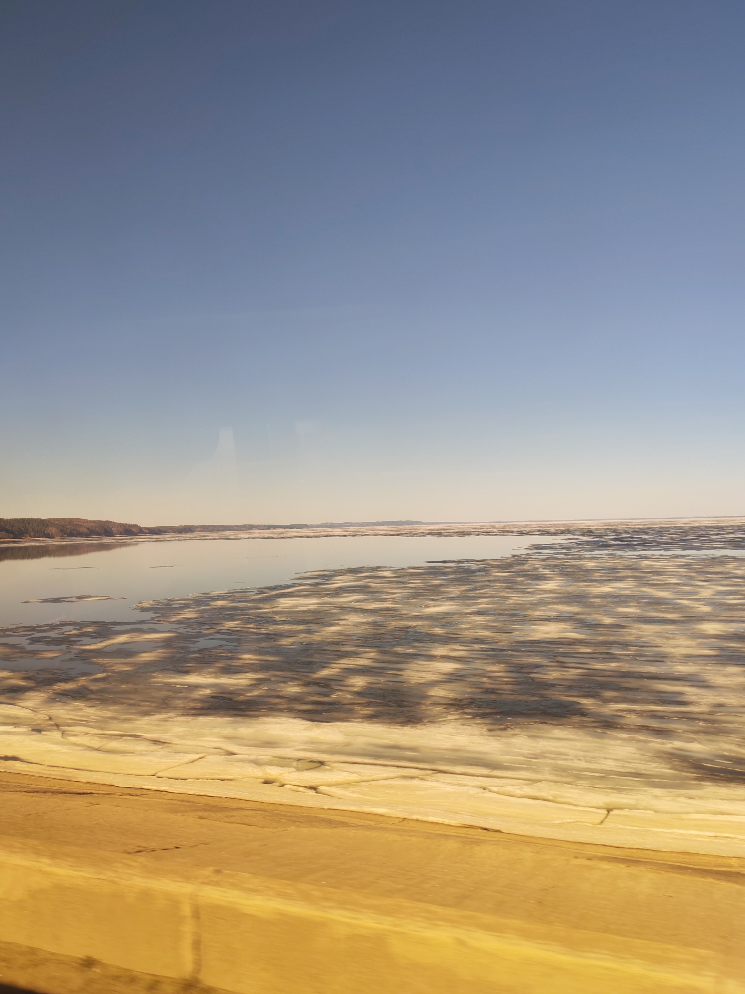 The image shows a body of water, partially covered with ice, under a clear blue sky. A shoreline with trees is visible in the distance. The foreground has a yellowish tint, possibly due to lighting conditions or the color of the surface from which the photo was taken. The scene likely depicts the Kama River in the city of Chaikovsky, Russia, based on the typical winter landscape of the region. No people or actions are discernible.