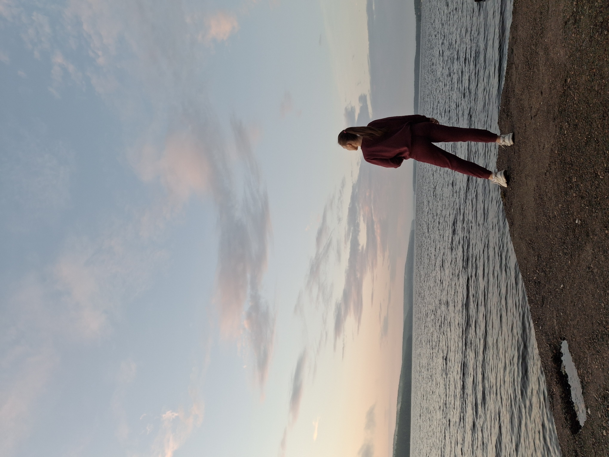 A young woman stands on a rocky shore facing a large body of water. She is wearing a maroon tracksuit and white sneakers. The sky is a pale blue with scattered pink and grey clouds, indicating either dawn or dusk. The water is calm, with gentle ripples reflecting the sky. In the distance, a hazy, forested shoreline can be seen. The location is likely a reservoir or large river, possibly the Kama River near Chaikovsky, Russia, given the characteristic landscape and flat horizon. The image is taken from a low angle, emphasizing the vastness of the water and sky.