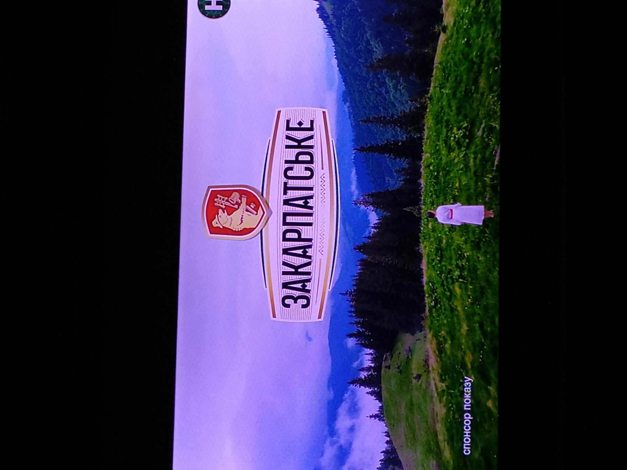 The image depicts a vertical view of a scenic mountain landscape with a logo and text. In the foreground, a white sign with a distinctive red and yellow shield logo is prominently displayed. The shield contains imagery of a bear and tools, possibly representing local heritage or industry. The sign itself has bold, vertical text in a foreign language, appearing to read "ЗAKAPПАТЬСЬКЕ" (Zakarpatske). Behind the sign, a woman in a white dress stands in a lush green meadow on a hillside. The background features a dense forest of dark green pine trees cascading down a mountain slope, with a hazy, purple-tinged sky and distant mountains visible. The scene suggests a natural, possibly rural or mountainous, setting. The lighting and muted colors indicate it could be late afternoon or early morning, with possibly overcast or misty weather. Towards the bottom right of the image, small, light-colored text reads "спонсор показу" which translates to "sponsor of the show" in Ukrainian.