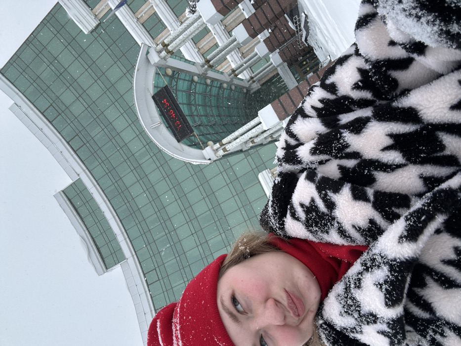 A young adult female is depicted in a partial frontal selfie taken outdoors during snowfall in Kazan, Russia. She occupies the lower-left foreground, wearing a red knitted beanie, a red turtleneck, and a black and white houndstooth-patterned faux fur jacket, all lightly dusted with snow. Her head is tilted slightly, and her lips are pursed in a subtle pout, with her gaze directed upwards and to the left. The background features the distinctive, curved green-tinted glass facade of the Kazan Family Center (Wedding Palace). A prominent entrance structure, partially obscured by the subject, includes an arched canopy under which a digital clock displays "13:46:54" in red characters. The surrounding environment is covered in snow, and individual snowflakes are visible throughout the frame, indicating active precipitation.