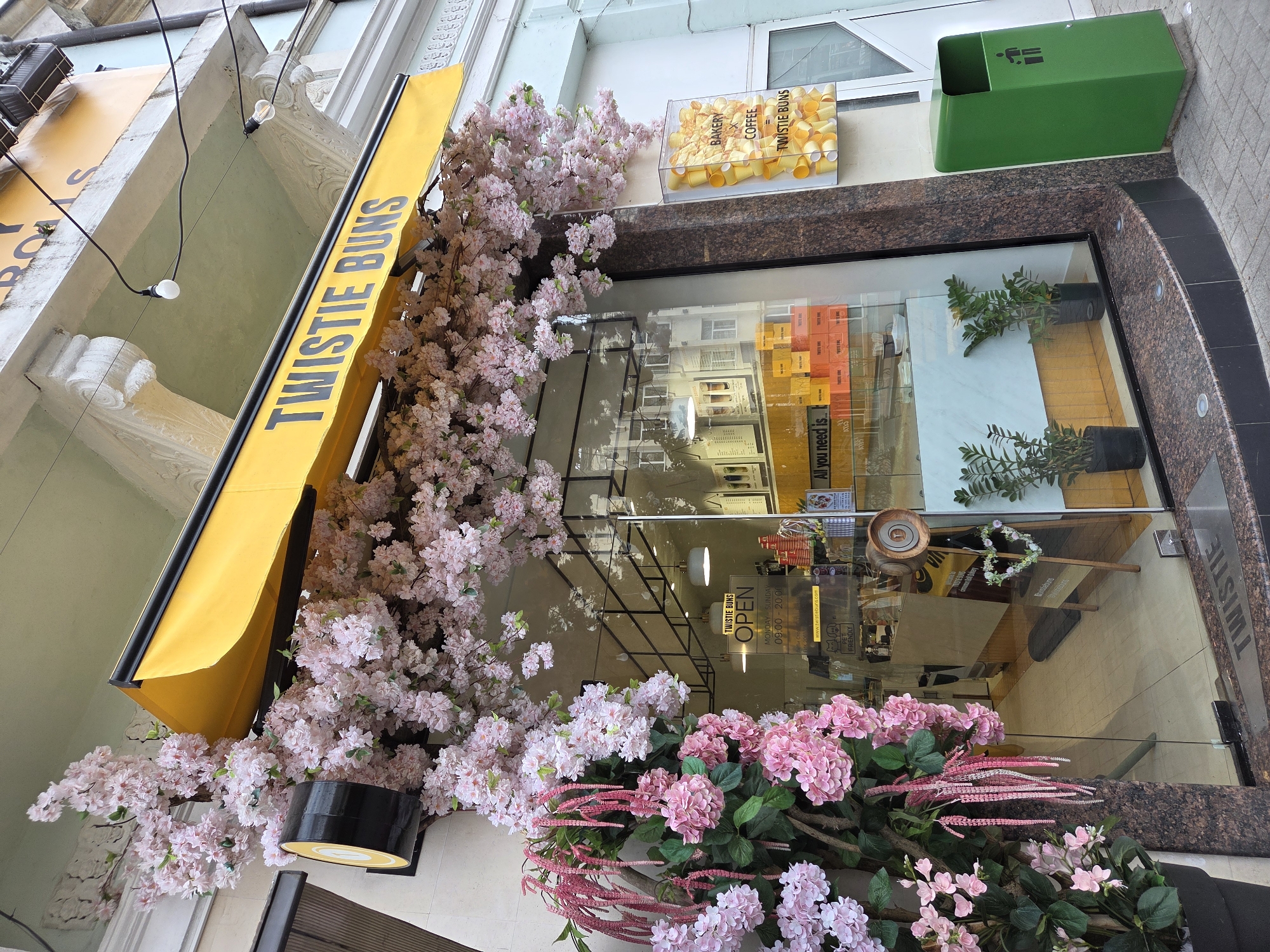 The image shows the facade of "Twistie Buns", a bakery and coffee shop. It has a yellow awning with "TWISTIE BUNS" written on it. Artificial pink flowers are attached to the facade and the awning. A transparent container on the wall displays yellow coffee cups with the brand's name on them. The window reflection shows a building opposite. A green trash bin is placed next to the shop's entrance. According to the information written on the window, the location is in Varna, Bulgaria.