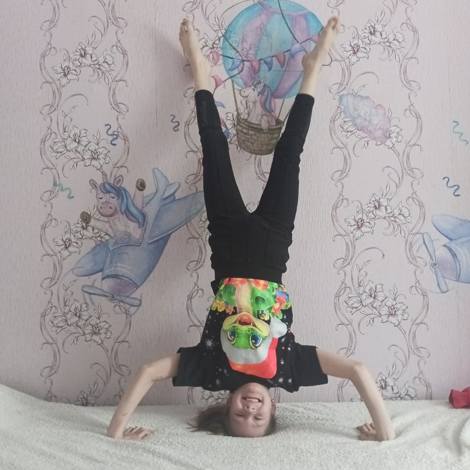 A young girl is upside down on a bed, striking a headstand pose. She is wearing black leggings, a black t-shirt with stars, and an article of clothing with an animated alligator or similar character around her mid-section. Her feet are in the air. She is smiling.
The background is a wall covered with wallpaper featuring images of unicorns in a hot air balloon and in an airplane, along with floral designs. The bed has a white, textured cover.
The image was taken in Chaikovsky, Russia.