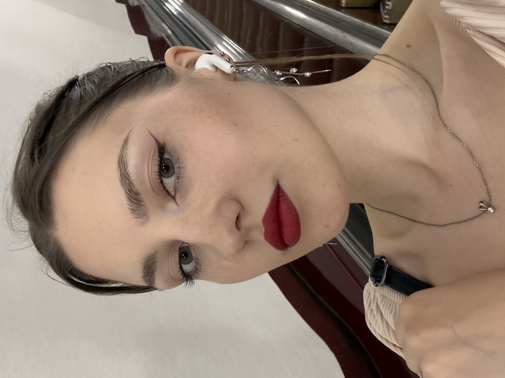 A young adult female is depicted in a close-up portrait within a Saint Petersburg Metro station, specifically on an escalator. She has fair skin, dark brown hair pulled back, and hazel eyes directly engaging the camera. Her makeup includes defined eyebrows, dark winged eyeliner, visible mascara on both upper and lower lashes, and matte red lipstick.
In her right ear, she wears a white Apple AirPods Pro earbud, positioned above a complex silver-colored dangle earring that features a swirling design and multiple clear faceted beads or crystals. Two small silver stud piercings are visible on the cartilage of her right ear. Around her neck is a delicate silver-colored chain necklace with a small, stylized animal pendant, possibly a rabbit, featuring a tiny central jewel. She is wearing a light-colored top, possibly beige or cream, with a visible ribbed or pleated texture on the shoulder. A black strap, likely from a shoulder bag, is visible over her left shoulder.
The background shows the characteristic architecture of a Saint Petersburg Metro escalator tunnel. On the right, the metallic handrail and steps of an upward-moving escalator are visible. Above the escalator, a distinctive curved paneling, appearing to be polished dark brown or maroon material, possibly wood or stone, ascends towards a lighter-colored upper wall or ceiling.