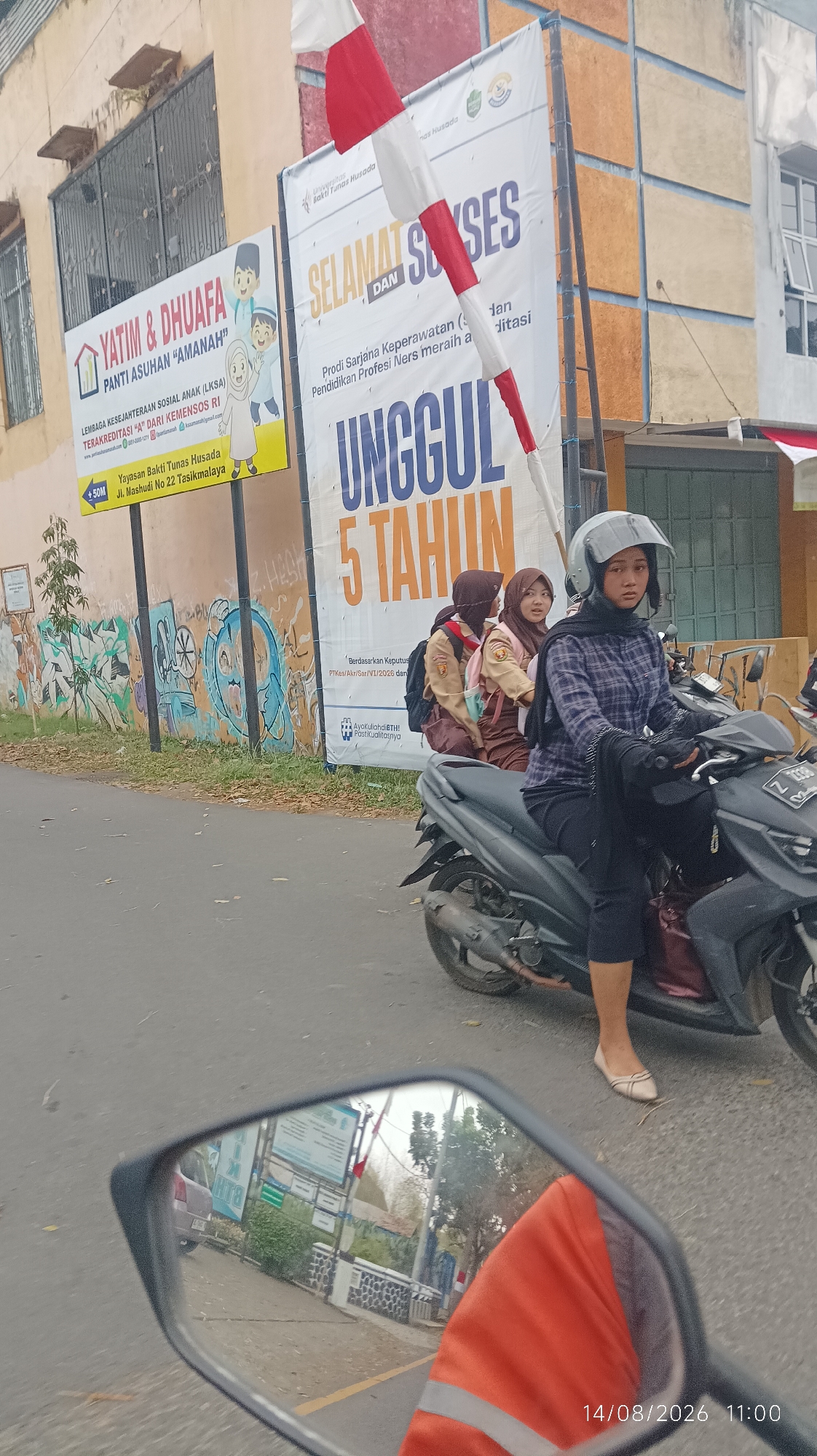 The image depicts a street scene in Tasikmalaya, Indonesia. In the foreground, a person wearing a helmet and a plaid shirt is riding a scooter, with two other individuals, presumably students in uniform, seated behind them. The scooter is on a paved road, with fallen leaves scattered on the roadside.
Behind the scooter, there are two large banners. The banner on the left advertises "YATIM & DHUAFA PANTI ASUHAN 'AMANAH'", an orphanage located at Jl. Mashudi No 22 Tasikmalaya. The banner on the right promotes "Universitas Bakti Tunas Husada", highlighting their "Prodi Sarjana Keperawatan (S)" and stating they have achieved "UNGGUL 5 TAHUN" (Excellent for 5 years). The building behind the banners appears to be a multi-story structure with colorful walls.
In the reflection of the scooter's rearview mirror, a building with a wall adorned with graffiti and a small tree can be seen. The license plate on the scooter is partially visible, showing "Z 3096". A timestamp at the bottom right of the image reads "14/08/2026 11:00".