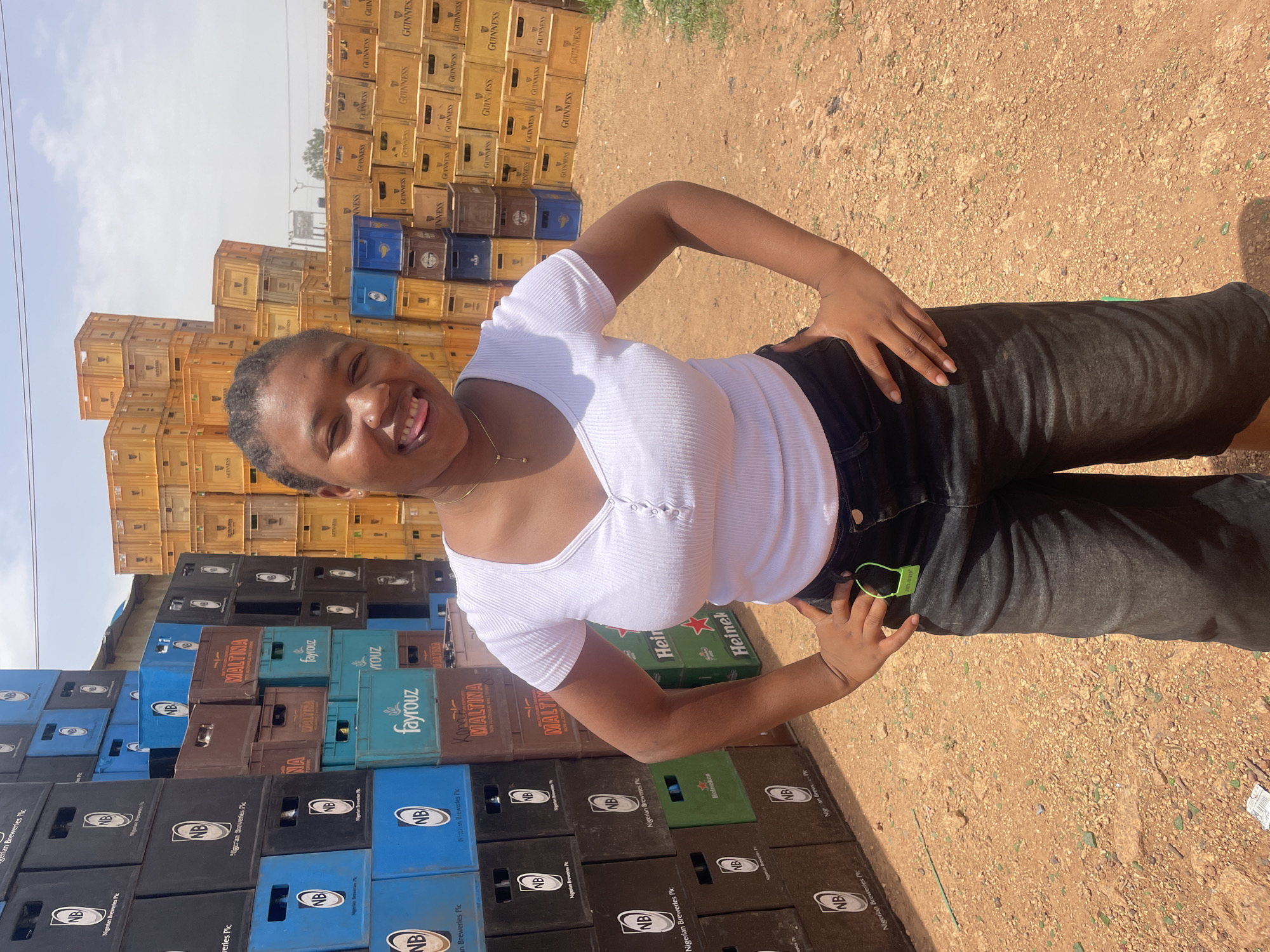 The image shows a woman in Gura Top, Nigeria, posing in front of stacks of stacked crates of beverages. The woman is wearing a white top and black pants. The crates are predominantly labeled with brand names such as Guinness, Heineken, Fayrouz, Maltina, and NB (Nigerian Breweries). The ground appears to be a reddish-brown dirt surface with small stones. The sky is visible in the upper left corner.
