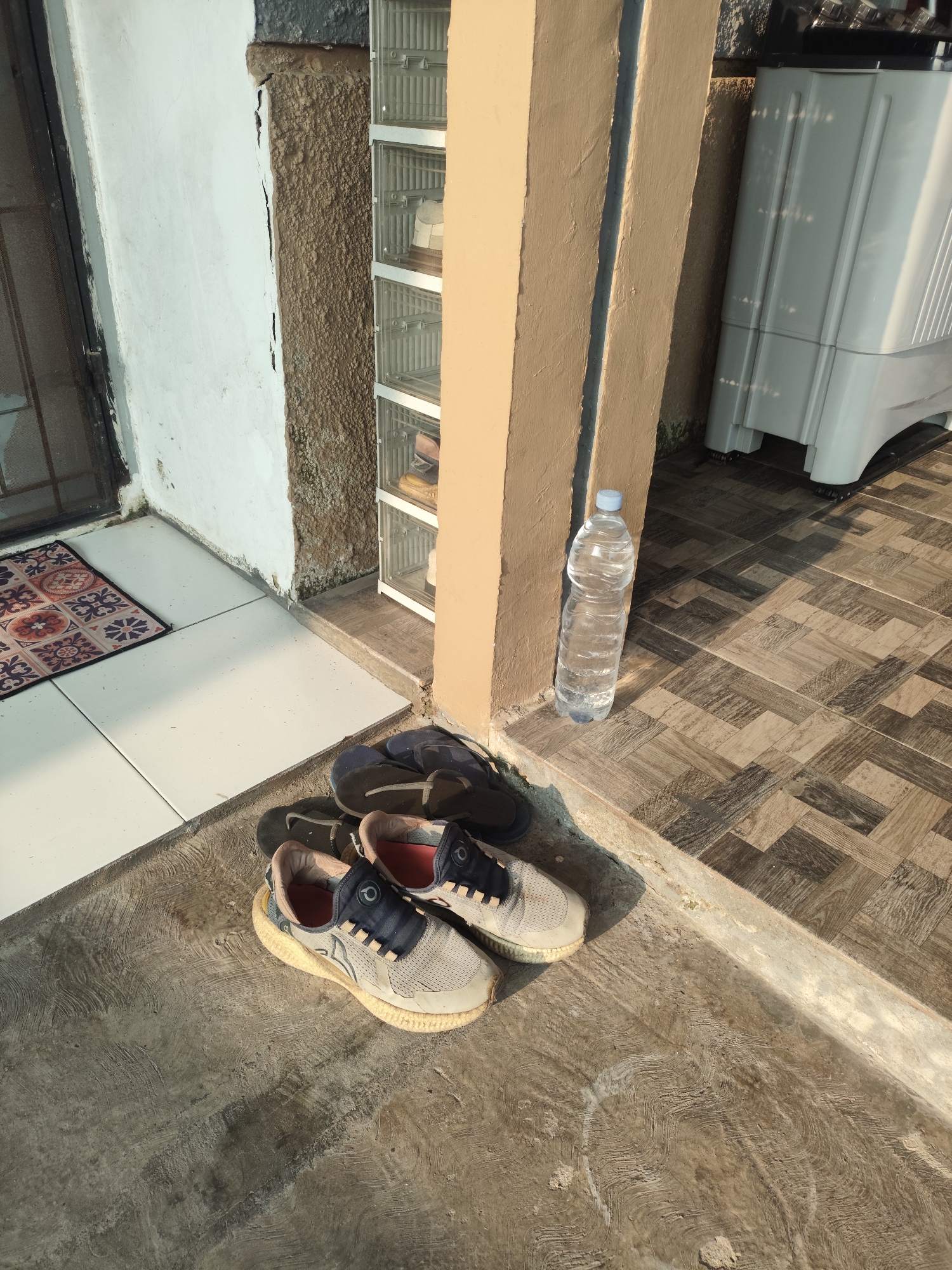 A pair of light-colored sneakers and a pair of dark flip-flops are placed on a concrete surface outside a building. Behind the shoes, a clear plastic bottle of water stands next to a pale yellow pillar. Further back, a white plastic washing machine is visible, partially obscured by the pillar. To the left, a white wall with a cracked section of rough texture meets a doorway with a patterned doormat on a tiled floor. The scene appears to be an exterior area, possibly a porch or entryway, located in Palembang, Indonesia, indicated by the characteristic architectural elements and common household items.
