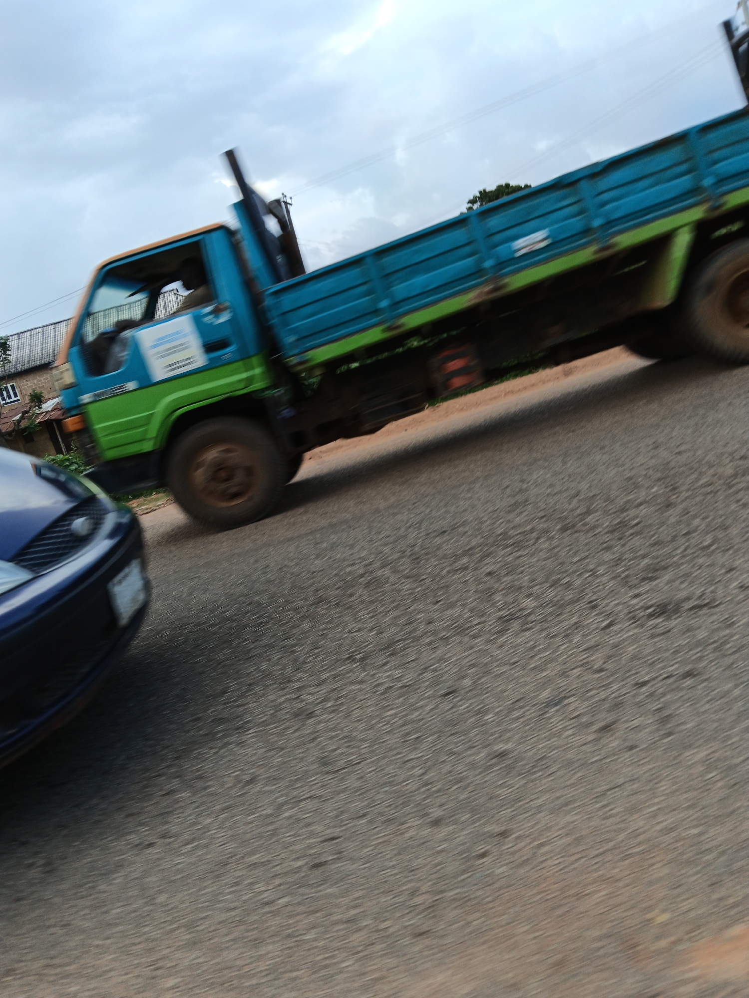 A blue and green flatbed truck is captured in motion, with a portion of a blue car visible in the foreground. The truck has a visible exhaust pipe and a turquoise colored bed with green trim. The driver is partially visible in the cab of the truck. The scene appears to be on a paved road in Vom, Nigeria, with some buildings and vegetation in the background. The sky is overcast with clouds. The image is slightly blurred due to movement.