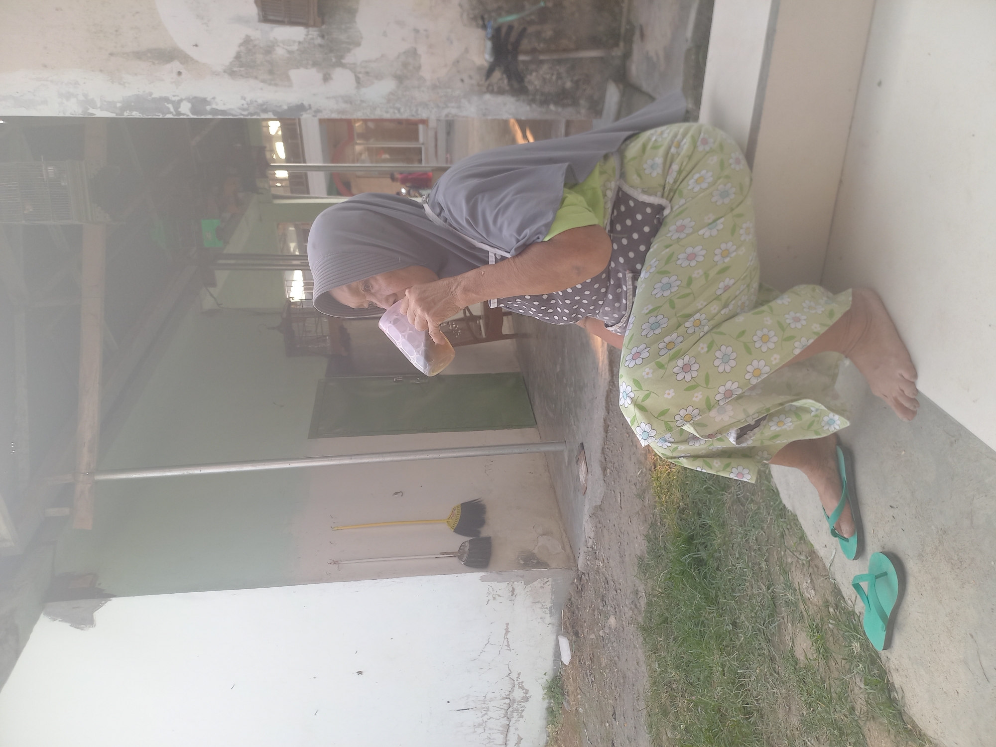An elderly woman wearing a grey hijab, a green t-shirt, a polka-dot apron, and floral patterned pants is sitting on a concrete step, drinking from a clear plastic cup. She is outdoors, with a building behind her and grass and dirt in the foreground. Two green flip-flops are on the ground near her feet. The building appears to be a house or small structure, with a light-colored exterior wall. Two brooms are leaning against the wall. In the background, birdcages hang from the roof. The scene is dimly lit, possibly due to overcast weather or the time of day.