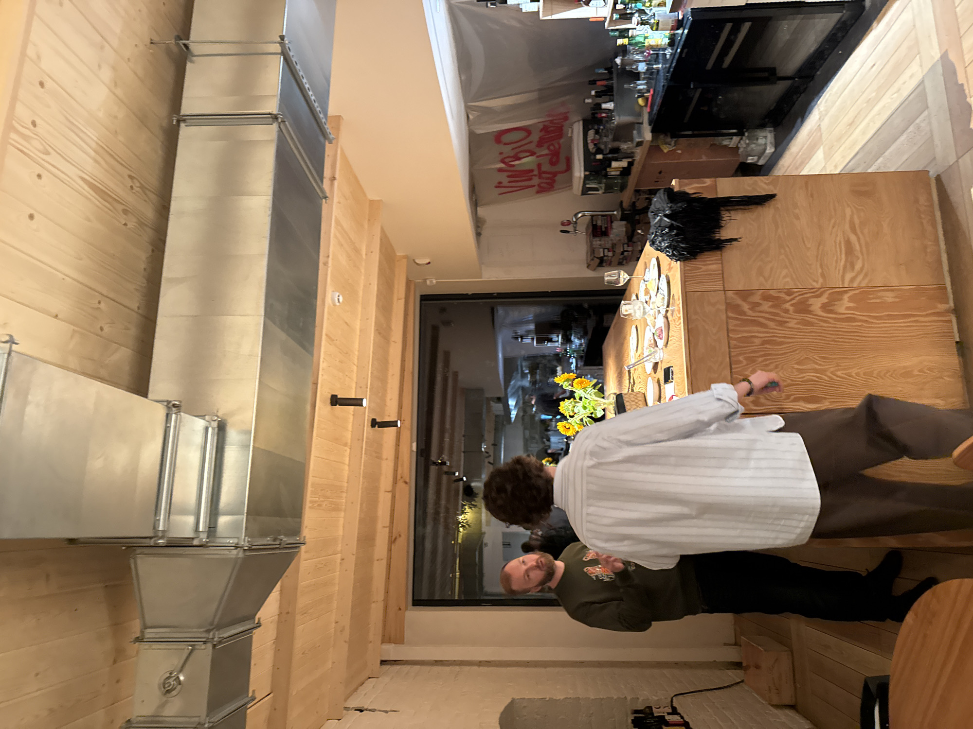 The image captures an interior scene at what appears to be a restaurant or bar, with a wooden ceiling featuring exposed ventilation ducts. Two men are the primary subjects. One man, facing left, has a beard and is wearing a dark green sweatshirt with an image on it. He is gesturing with his right hand. The other man, facing away from the camera, has curly brown hair and is wearing a light blue and white striped button-down shirt and brown pants. He is looking towards the first man.
In the foreground, a long wooden counter or island is visible, set with plates, glassware, and a vase of sunflowers. Behind the counter, a bar area is equipped with bottles of liquor and other beverages, as well as what looks like a beer tap. A white sheet or tarp hangs on the wall behind the bar, with red graffiti on it. In the background, through a large glass partition, the interior of another section of the establishment is dimly lit, showing kitchen equipment and more people.
The scene is indoors, and the lighting suggests it is evening. The overall aesthetic is modern and rustic, with natural wood and industrial elements. The graffiti on the wall reads "VIN BIO" followed by other illegible text. The flooring is light-colored wood planks. The atmosphere appears to be social, with the two men engaged in conversation. The location is identified as Moscow, Russia.