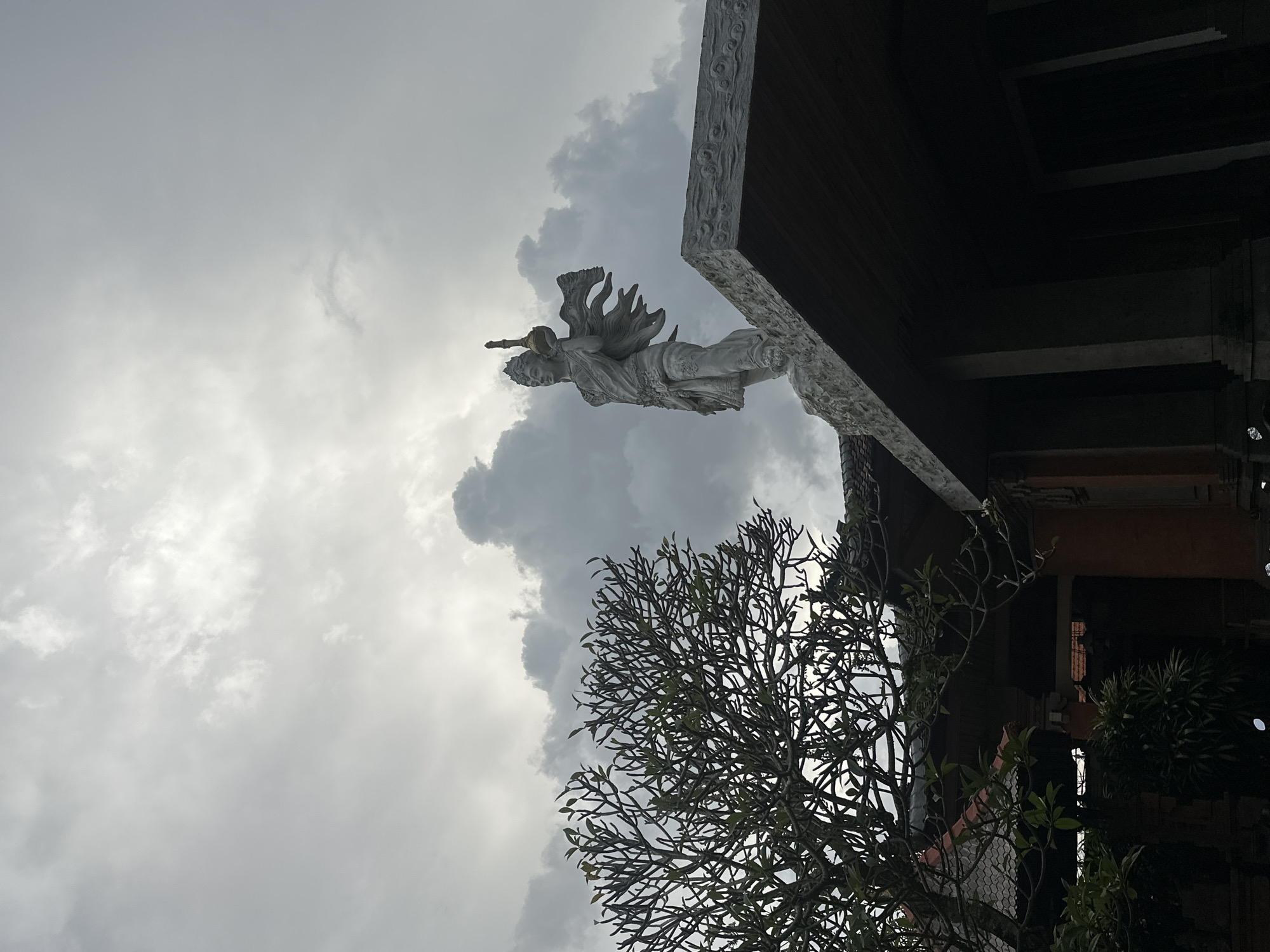 The image shows a cloudy sky above a building in Celuk, Indonesia. A stone statue of a person with ornate wings and an instrument is fixed atop the building's edge. A tree with a dense network of branches is visible below. The building is made of dark wood, with intricate carvings along the edge.
