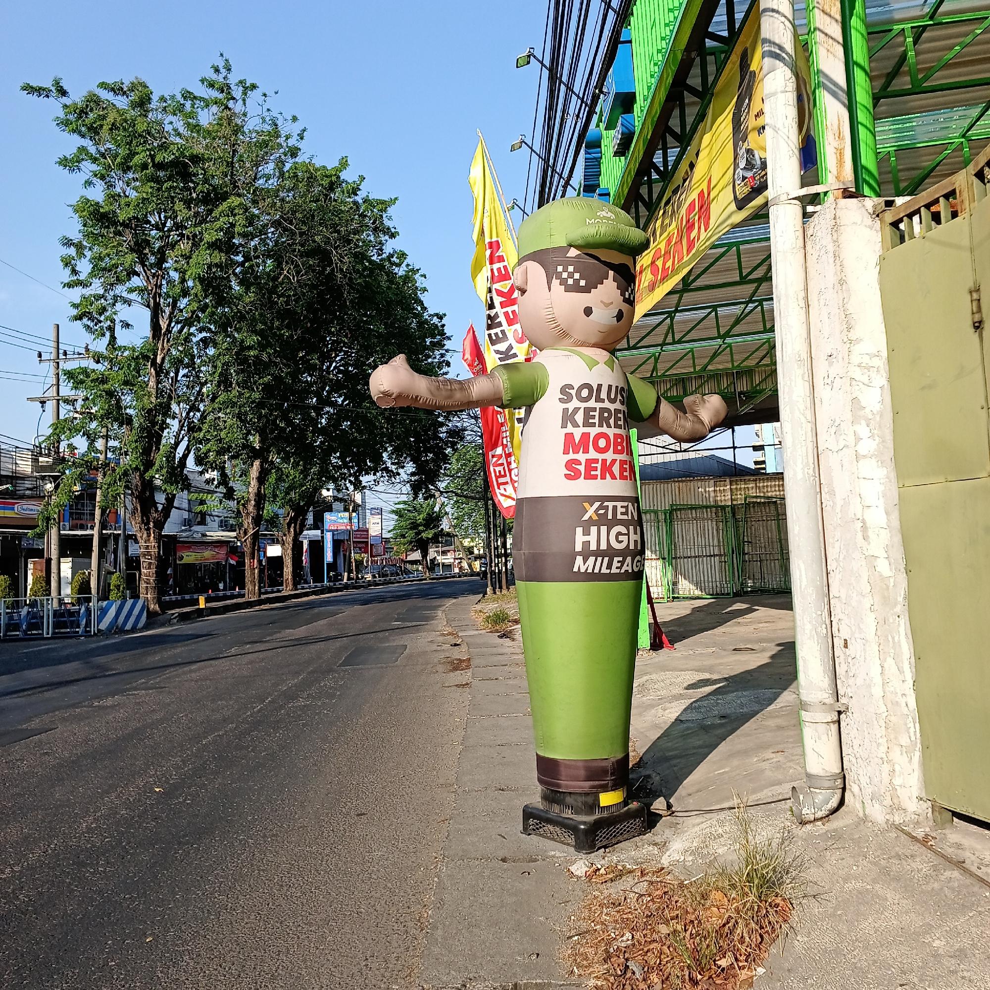 An inflatable figure stands on the sidewalk in front of a building with a green awning. The figure is dressed in a green cap and shirt with the words "SOLUSI KEREN MOBIL SEKEN" and "X-TEN HIGH MILEAGE" printed on it. Two flags, one yellow and one red, are planted behind the inflatable figure. The building appears to be a business, with signs and awnings visible. The street in front of the building is paved and shows signs of wear. Trees line the sidewalk, and there are power lines overhead. The scene is lit by bright sunlight, suggesting it is daytime. The location is identified as Wedoro, Indonesia.