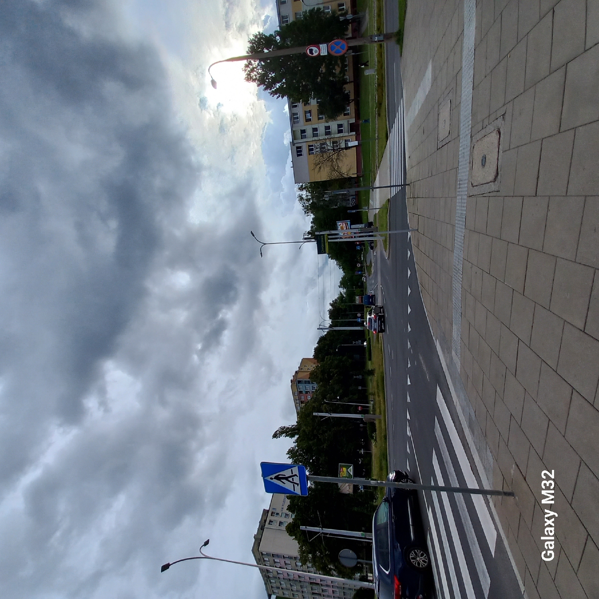 The image depicts an urban street scene in Olsztyn, Poland, on an overcast day. In the foreground, a pedestrian crossing with white diagonal stripes is visible on the asphalt road, bordered by a sidewalk paved with grey square tiles. A dark blue SUV is parked to the left, partially obscuring the pedestrian crossing. A signpost with a blue square sign displaying a white pedestrian silhouette is planted on the sidewalk. Beyond the road, several apartment buildings with multiple stories and numerous windows are situated, interspersed with a dense cluster of green trees. Streetlights with curved arms extend over the roadway. In the distance, other vehicles are visible on the road, and more buildings and trees populate the landscape. The sky is filled with heavy, grey clouds, with a sliver of bright sky peeking through on the right side, suggesting the sun might be present behind the clouds. The "Galaxy M32" watermark is present in the bottom left corner of the image.