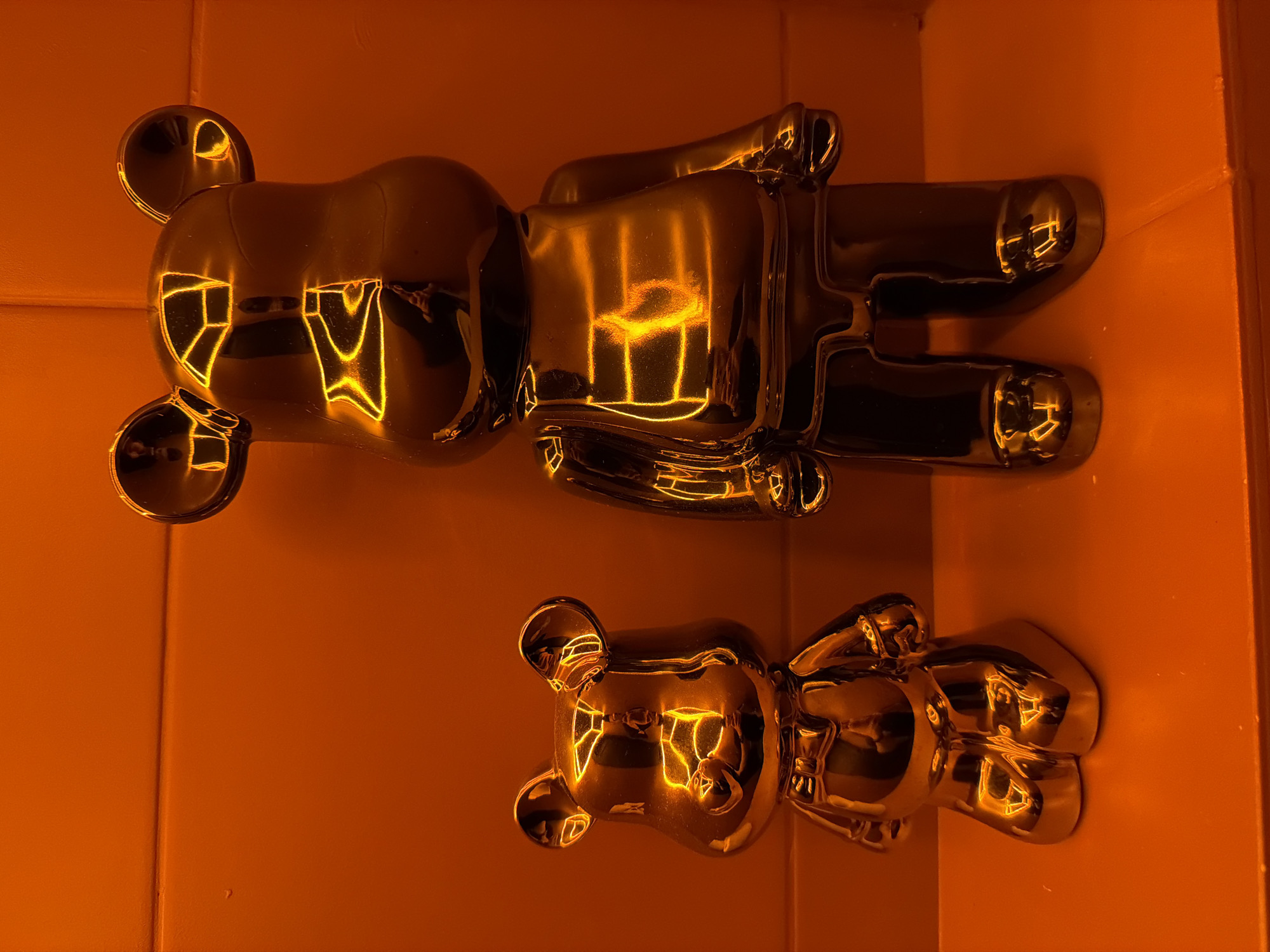 Two gold-colored bear-shaped figures are displayed against an orange background. The larger figure lies horizontally at the top of the image, while the smaller figure is positioned beneath it. Both figures have a reflective surface that captures light. There is no indication of a specific location in Izhevsk, Russia, or any people or actions in the scene.