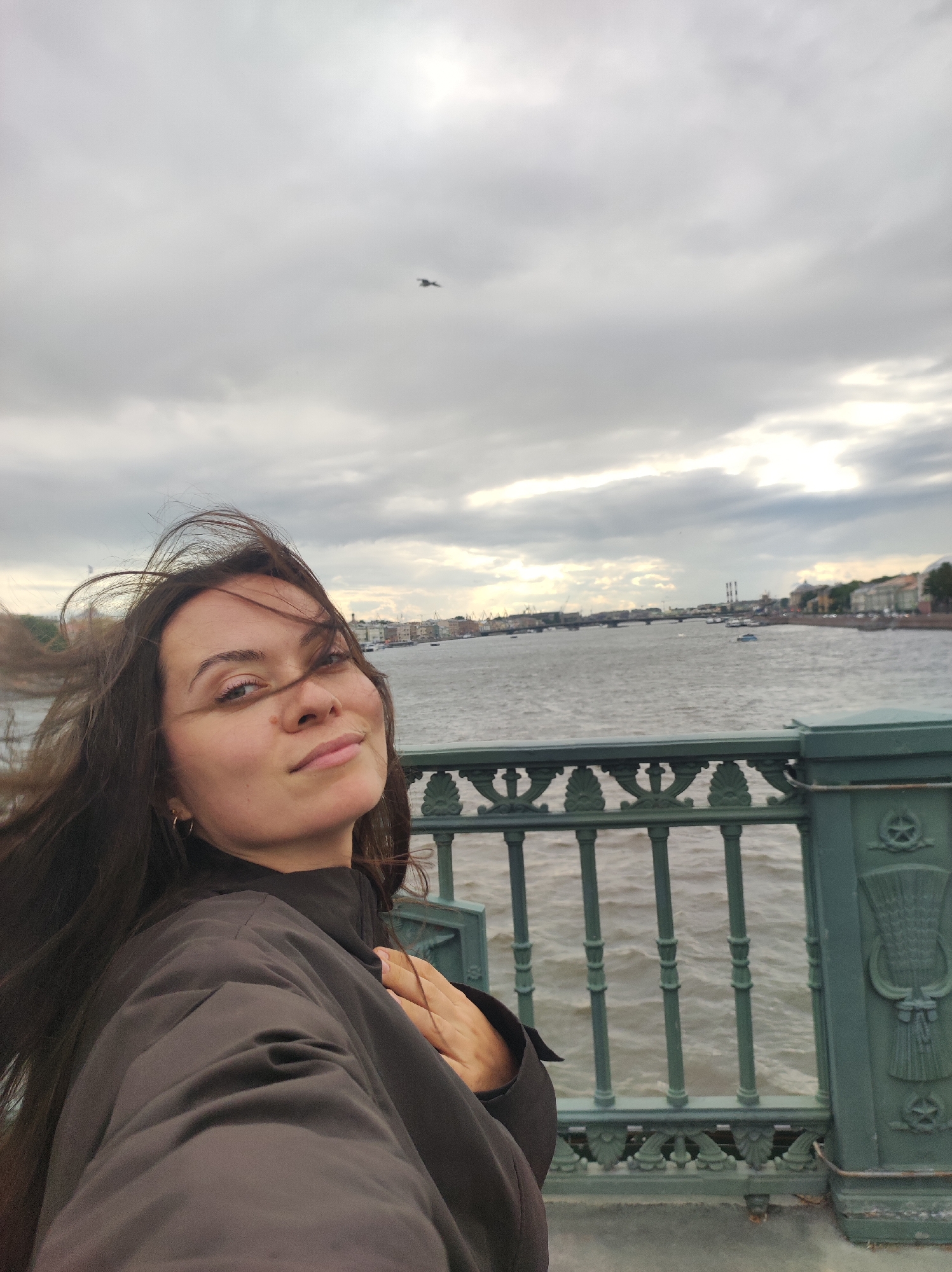 A woman with long, dark hair is taking a selfie on a bridge. She is wearing a dark jacket and has her left hand on her chest. Her hair is being blown by the wind. Behind her, a body of water is visible, with boats and buildings on the opposite bank. The sky is overcast. The bridge has a green, ornate railing. A bird is flying in the sky above the water.