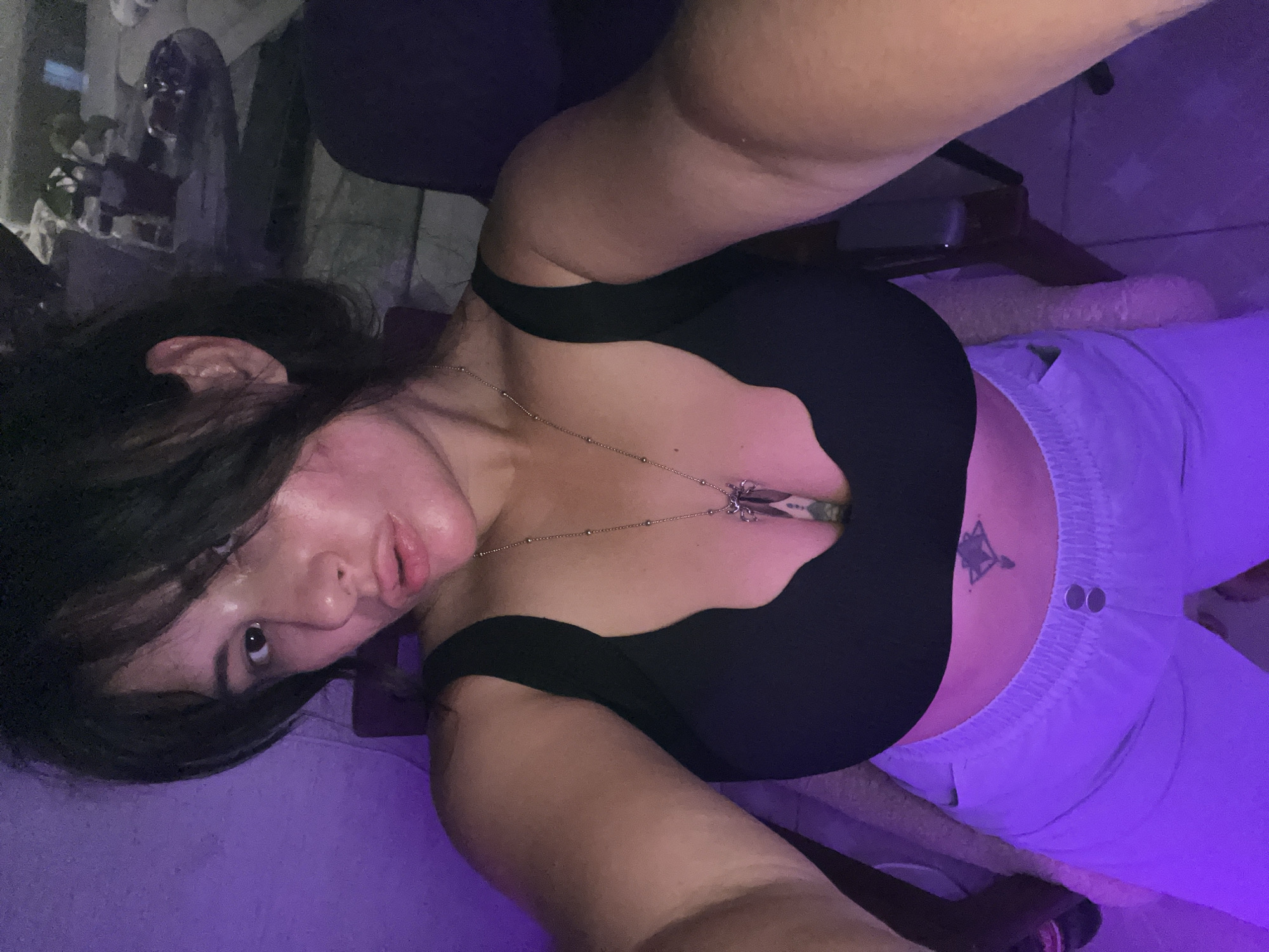 In Kiryat Bialik, Israel, a woman is taking a selfie. She is seated and wearing a black sports bra and light-colored sweatpants. A silver necklace with a pendant hangs around her neck. The background is dimly lit, with purple lighting casting a glow on the scene. The woman's hair is dark and falls around her shoulders. She has a tattoo on her abdomen.