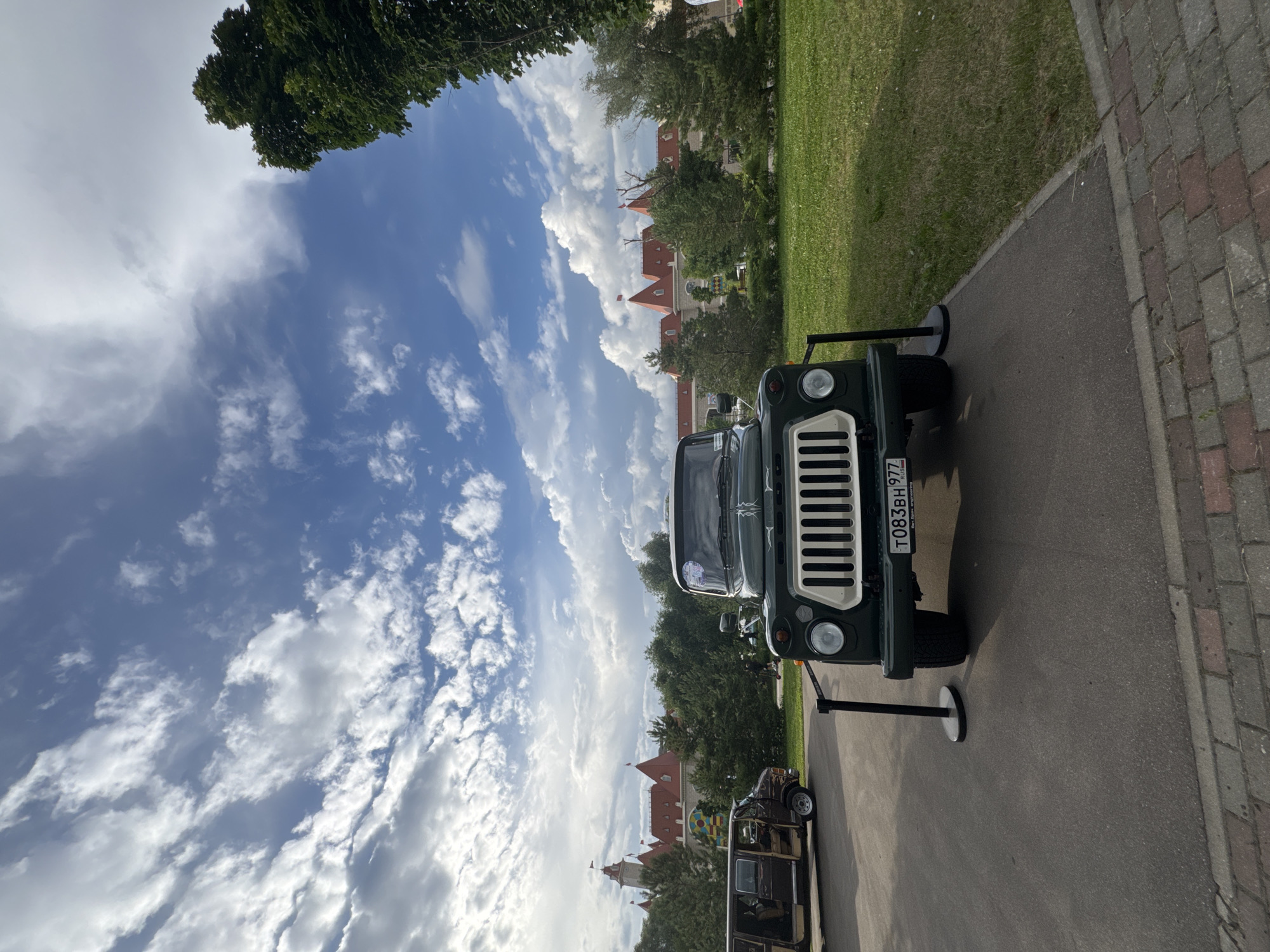 This image captures a sunny day in Moscow, Russia. The scene features a vintage dark green vehicle with a white grille parked on an asphalt path. The vehicle has a license plate that reads "T083BH 977". Behind the vehicle, there is a lush green lawn, trees, and a building with red-tiled roofs, possibly a historic castle or palace. The sky is a vibrant blue with fluffy white clouds, suggesting pleasant weather. Another older vehicle, possibly a van, is visible in the background. The overall impression is a picturesque outdoor setting with vintage vehicles displayed against a backdrop of historic architecture. The time of day appears to be midday, given the bright sunlight casting shadows.