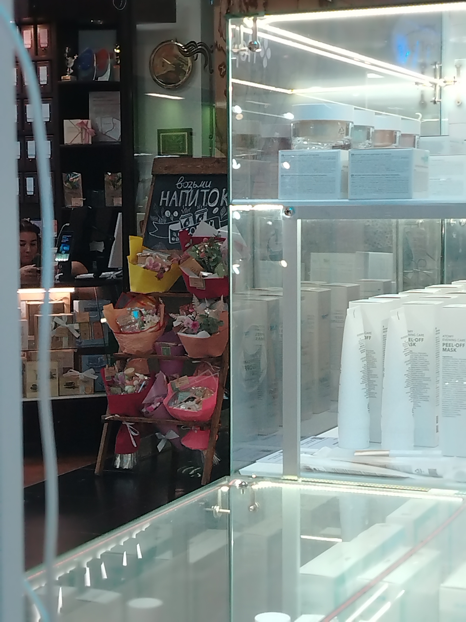 The image shows a shop in Izhevsk, Russia, featuring a display case filled with cosmetics and skincare products, including tubes labeled "ATOMY EVENING CARE PEEL-OFF MASK". Outside the display case, there is a blackboard advertising "drinks" (напиток), along with colorful bouquets and wrapped gifts. A woman is partially visible behind the counter. A clock hangs on the wall in the background, adding to the scene's environment.