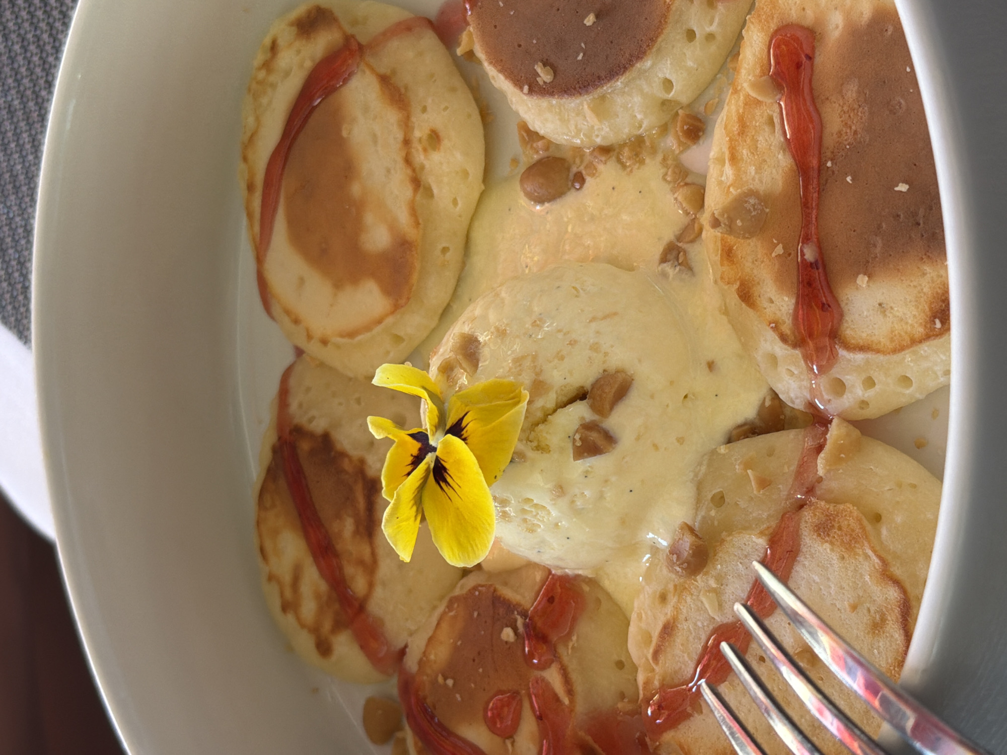 The image shows a close-up of a dessert in a white bowl, likely at a restaurant in Budva, Montenegro. The dessert consists of several small, round pancakes drizzled with a red sauce, possibly strawberry or raspberry coulis. In the center, a scoop of pale yellow ice cream is adorned with a vibrant yellow edible flower. Scattered around the ice cream and pancakes are small, brown, crunchy pieces, which appear to be nuts or toffee bits. A silver fork is partially visible on the left side of the frame, positioned over some of the pancakes. The background is blurred, suggesting an indoor setting with ambient lighting.