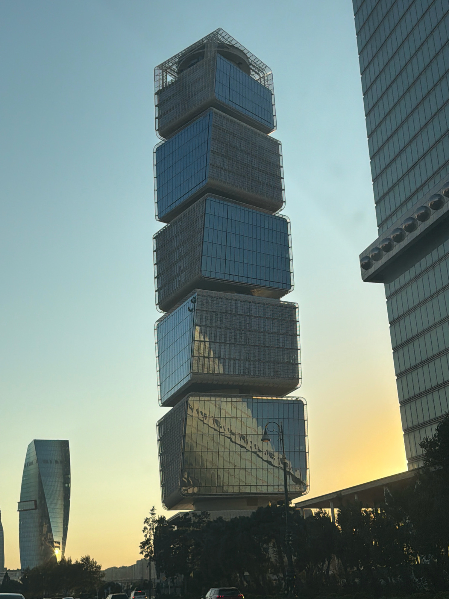 The image depicts a cityscape in Baku, Azerbaijan, at sunset. The foreground is dominated by a uniquely designed skyscraper composed of stacked, cuboid sections, each featuring a reflective glass facade. To the right of this building is another, more conventional skyscraper with a grid-like exterior. In the background, a distinctively twisted, glass-clad building stands out, reflecting the warm hues of the setting sun. The sky transitions from a clear blue at the top to a soft yellow near the horizon, indicating the late afternoon or early evening. Silhouetted trees and a street lamp add depth to the scene, and the presence of cars suggests an urban environment. The overall impression is one of modern architecture against a natural, atmospheric backdrop.