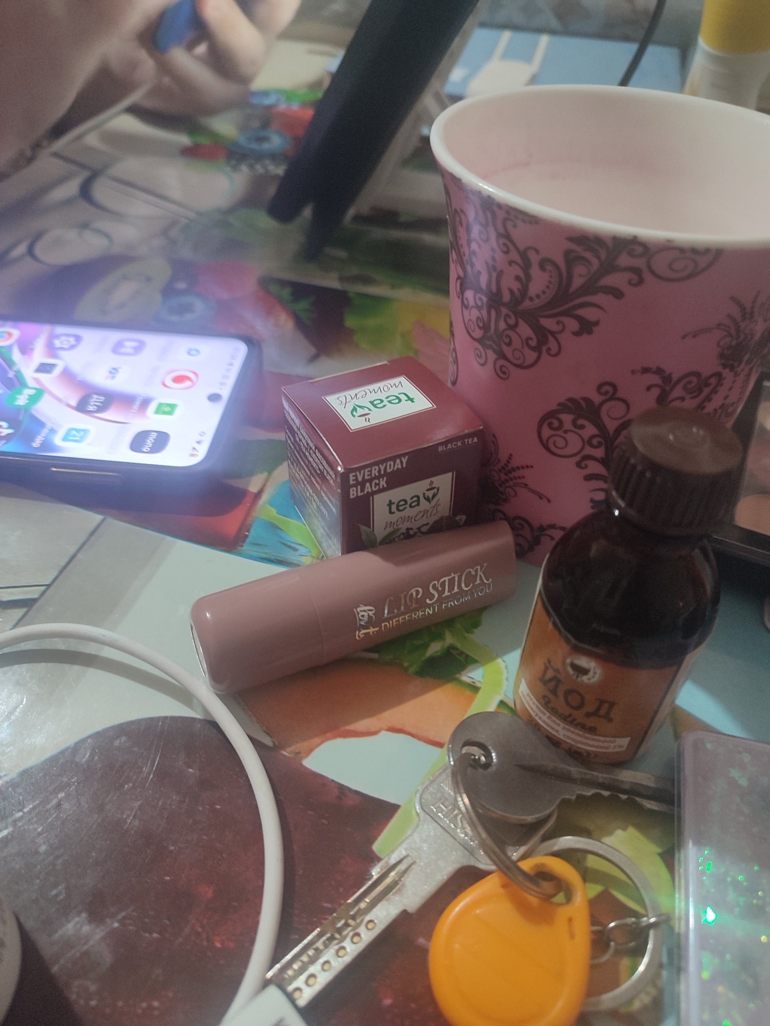 The image depicts a close-up, overhead view of a table surface, likely in a domestic setting in Chernivtsi, Ukraine. A smartphone with a lit screen, displaying various app icons including a phone, calendar, and messages, is positioned on the left. Adjacent to the phone is a small, dark red box with text that reads "TEA MOMENTS BLACK TEA." To the right of the tea box, a pink paper cup with a black, ornate design contains a pale pink liquid. In front of the tea box, a tube of lipstick with the text "LIP STICK" is visible. Further to the right, a small brown glass bottle with a dark cap contains a light brown liquid and has a label with Cyrillic text that appears to be "ЙОД" (IODINE). A set of keys, including a silver key and a yellow key fob, are resting on the table near the bottle. A white charging cable is also present, partially obscuring the bottom left of the frame. The background shows a patterned surface, possibly a tablecloth or placemat, with muted colors and floral or abstract designs. A person's hands are partially visible in the upper left corner, holding a blue object. The overall lighting suggests an indoor environment, possibly with artificial light.