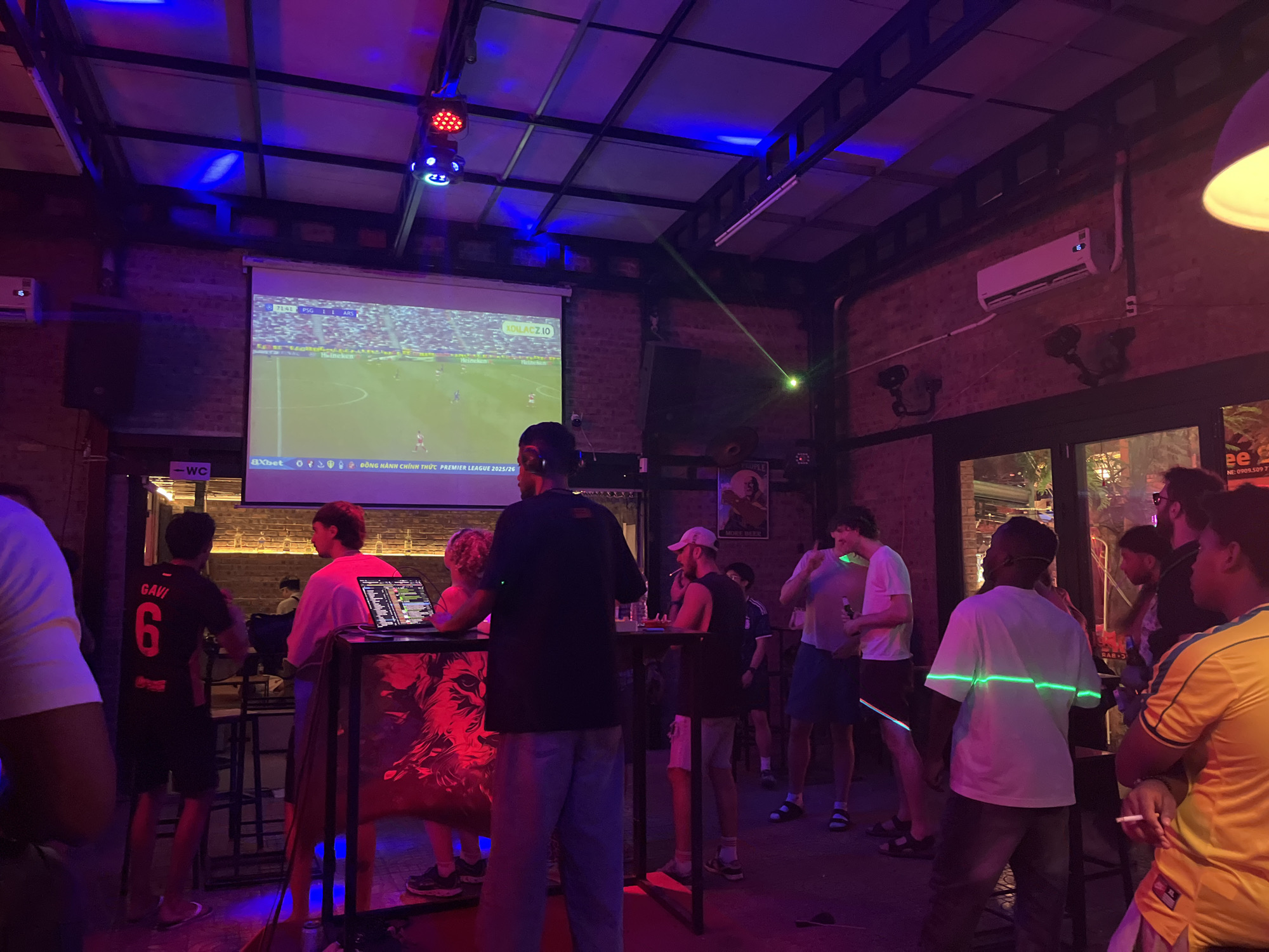 The image shows the interior of a bar in Đà Nẵng, Vietnam. A soccer match is being projected onto a screen, with a visible score of PSG 1 - 1 ARS at the 71:41 mark. Numerous people are standing and watching the game. There is a DJ booth set up with equipment. The lighting is predominantly blue and red. A green laser beam cuts across the room. There is an air conditioning unit mounted on the wall. Signage indicating "WC" (water closet) is visible. Various beer advertisements are present. Several patrons are holding or smoking cigarettes. One individual is wearing a soccer jersey with the name "GAVI" and the number "6" on the back. A sign includes the Vietnamese phone number "NE: 0909.5097".
