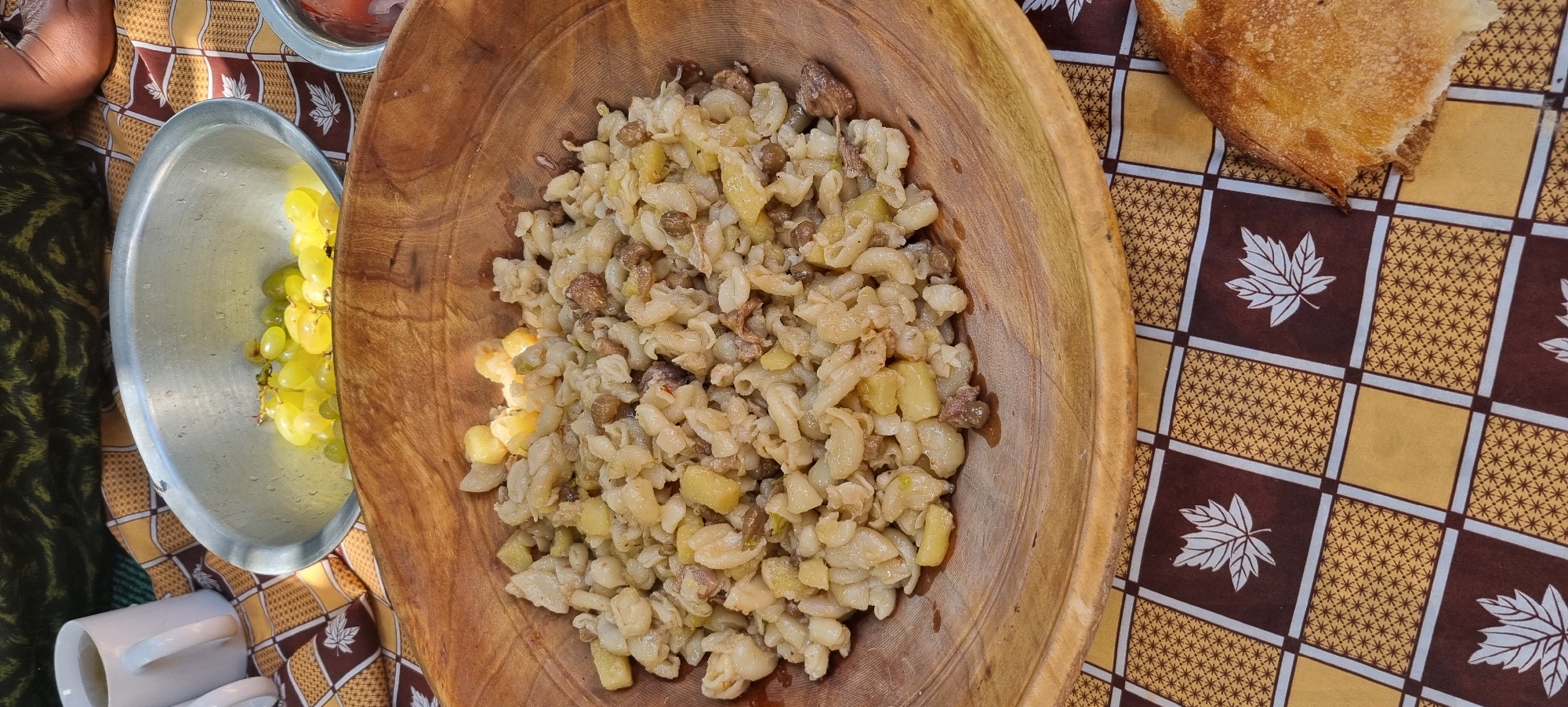 An overhead shot shows a close-up of a wooden bowl filled with a hearty mixture of what appears to be pasta, potatoes, and meat, seasoned and cooked together. The dish has a slightly greasy sheen, indicating it might be freshly prepared or served warm. The bowl itself is rustic, with a visible wood grain. It sits on a tablecloth with a checkered pattern of brown and gold squares, each adorned with a white leaf motif. To the left of the bowl, a metal bowl is partially visible, containing a cluster of light green grapes. A white mug with a handle is also visible to the upper left, and a slice of crusty bread with a golden-brown crust is positioned in the lower right corner of the frame, resting on the tablecloth. The overall scene suggests an outdoor or informal meal setting, possibly a picnic. The lighting is natural and appears to be daylight, casting soft shadows. The location is identified as Jomi, Tajikistan.