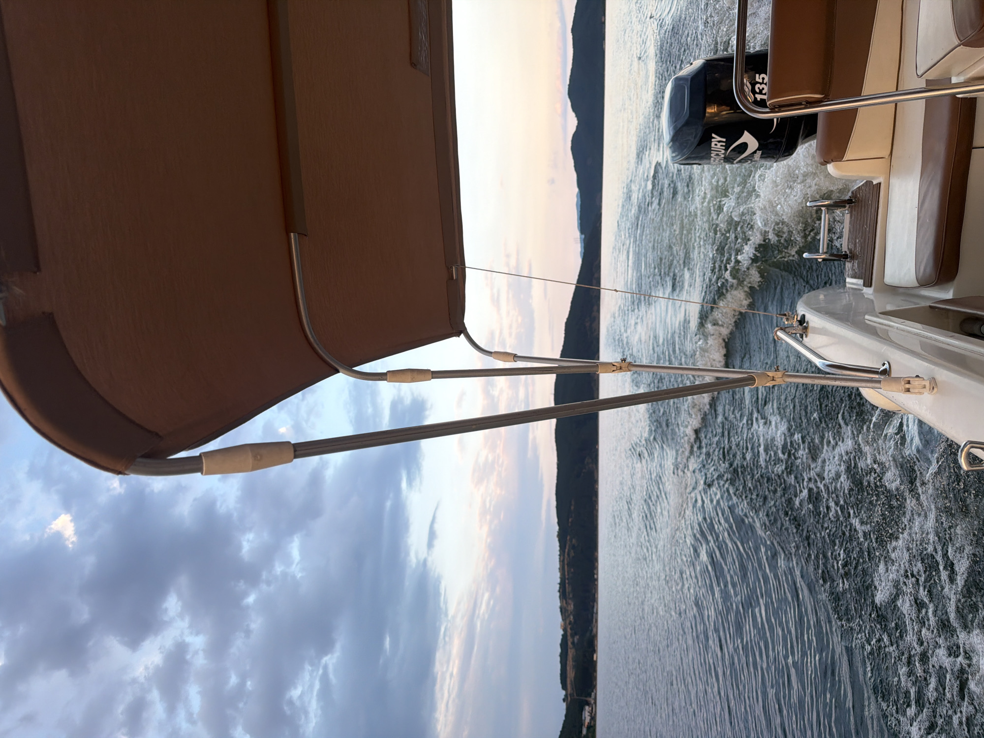 This is a perspective shot from a boat moving on the body of water of Glavatartsi, Bulgaria. The boat is traveling away from the camera, creating a wake behind it. The foreground features the back of the boat, including a brown and white upholstered seat, metal railings, and the boat's outboard motor, a black Mercury with "135" visible. The middle ground is dominated by the dark blue water with white foamy waves from the wake. The background shows a distant, tree-covered shoreline with a few buildings visible on a gentle slope. The sky above is filled with dramatic clouds, transitioning from a lighter shade near the horizon to darker grays and blues higher up, with hints of pink from what appears to be dusk.