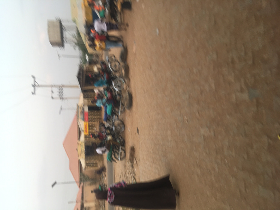 This medium-shot image captures a bustling street scene in Pindiga, Nigeria, likely during daytime under an overcast sky. The foreground and much of the right side are dominated by a wide, unpaved, reddish-brown dirt road or ground.

In the midground, numerous people are gathered alongside a row of single-story buildings. A significant number of motorcycles are parked or clustered around the individuals, suggesting a local transport hub or marketplace. The people, mostly appearing to be men, are indistinct due to blur and distance, but seem to be standing, conversing, or waiting. One person in the far foreground, on the right, is dressed in dark, flowing attire.

The buildings lining the road are simple in construction, likely concrete or brick, with prominent corrugated metal roofs. One particular building features a bright yellow facade and clearly displays a red and yellow "DHL" logo, indicating a delivery service point.

In the background, on the left, a large, square water tank is elevated on a tall metal stand, and several utility poles with power lines crisscross the sky. The dusty ground and the concentration of motorcycles and people suggest a dry climate and a vibrant, communal atmosphere typical of a semi-urban or rural area in Nigeria.